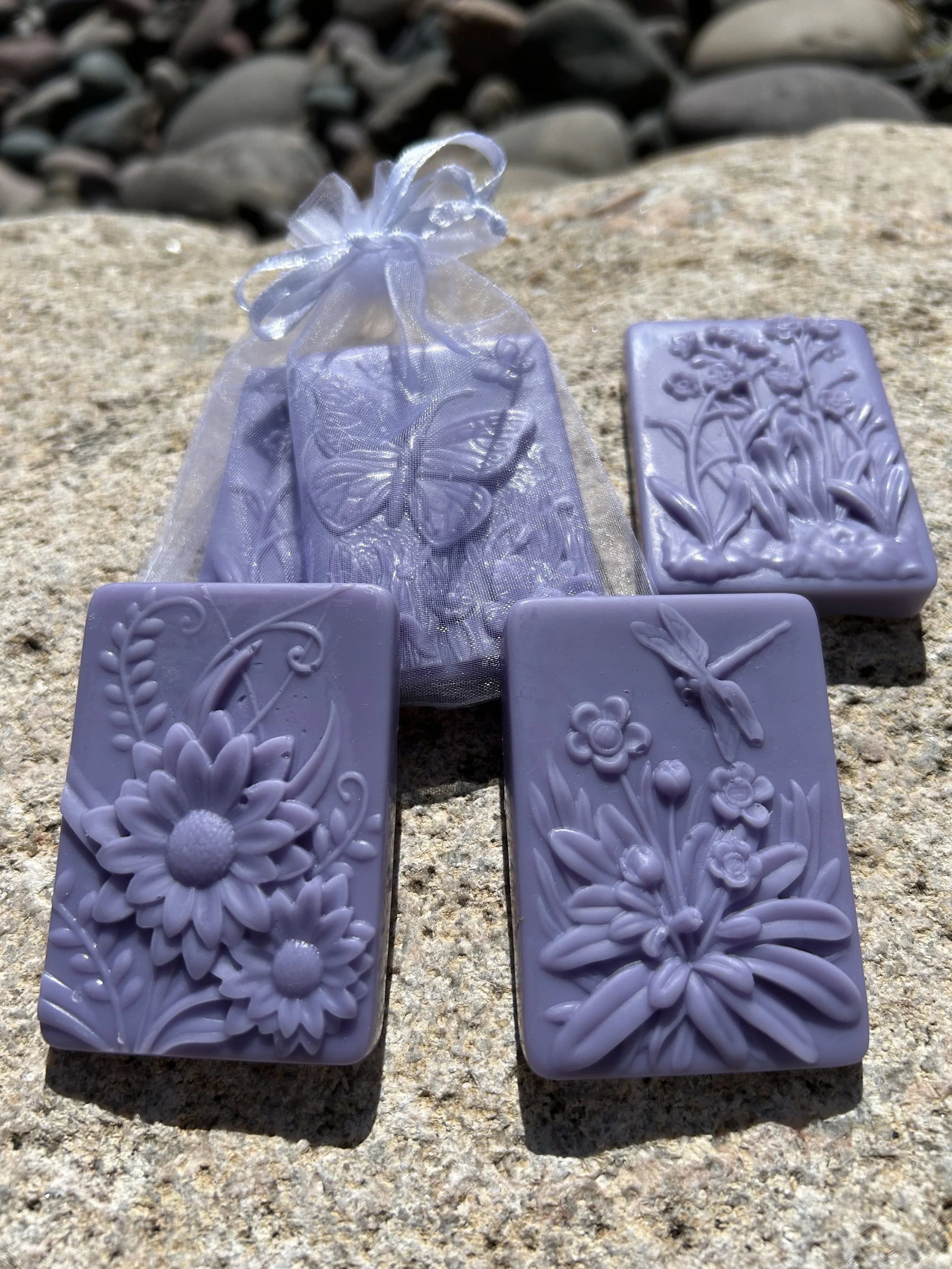 Purple lavender wax melts in the shape of bars with different floral and butterfly designs on them. They are placed for display on a rock outdoors.