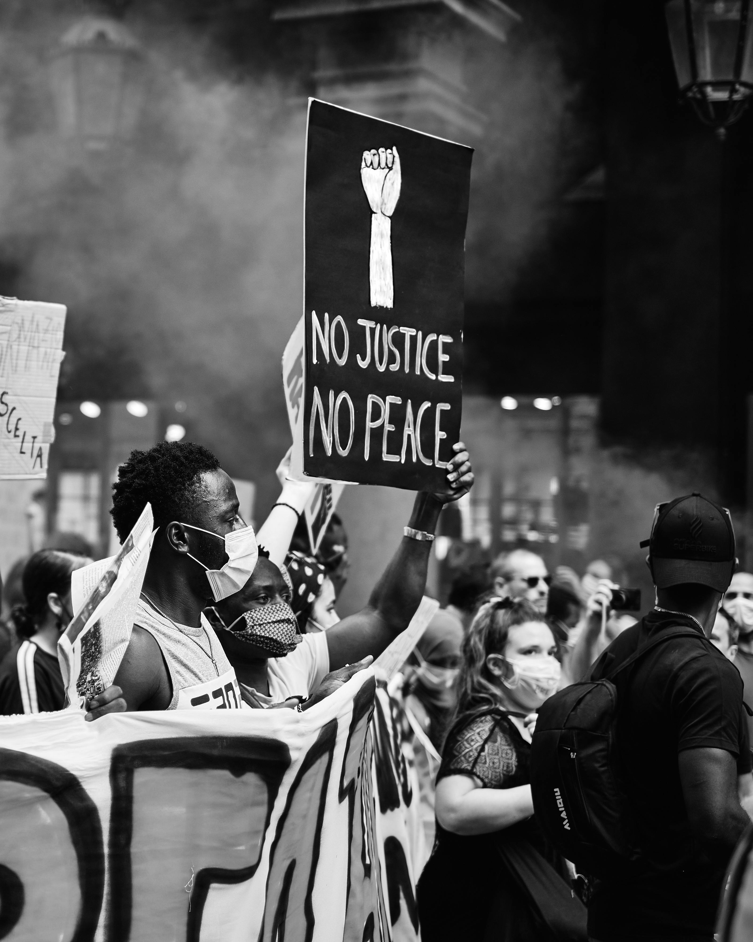 A black and white photo of a protest with people wearing masks. One protester holds a sign with a clenched fist and the words 'No Justice No Peace'.