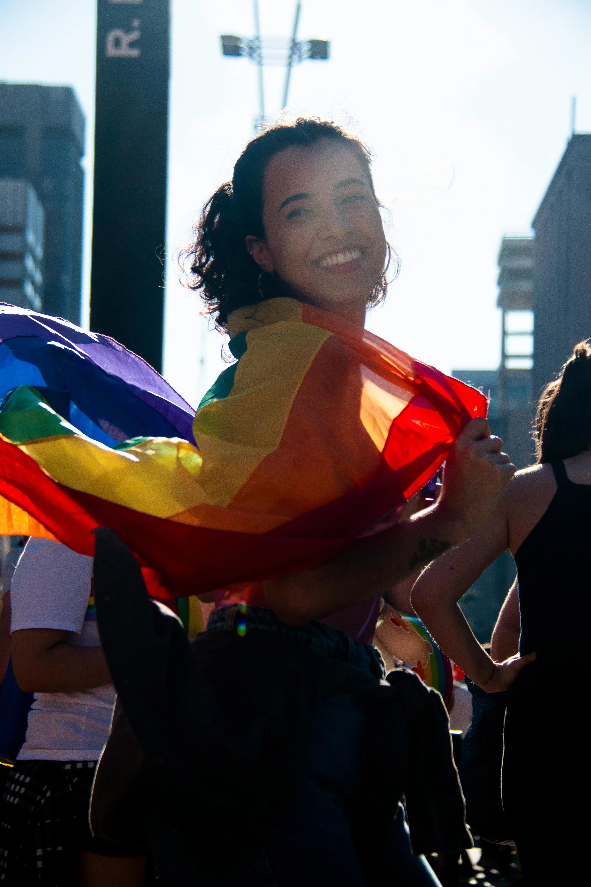 LGBTQ+ activist smiling and holding a rainbow pride flag during a parade, with a bright sunlit urban background.
