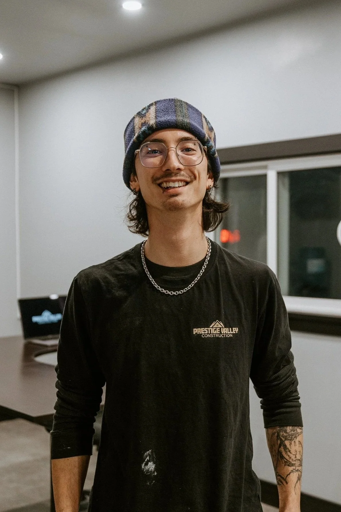 A smiling young man with glasses, a beanie, a chain necklace, earrings, and tattoos on his left arm, standing indoors in a room with a window and a desk with a computer in the background.