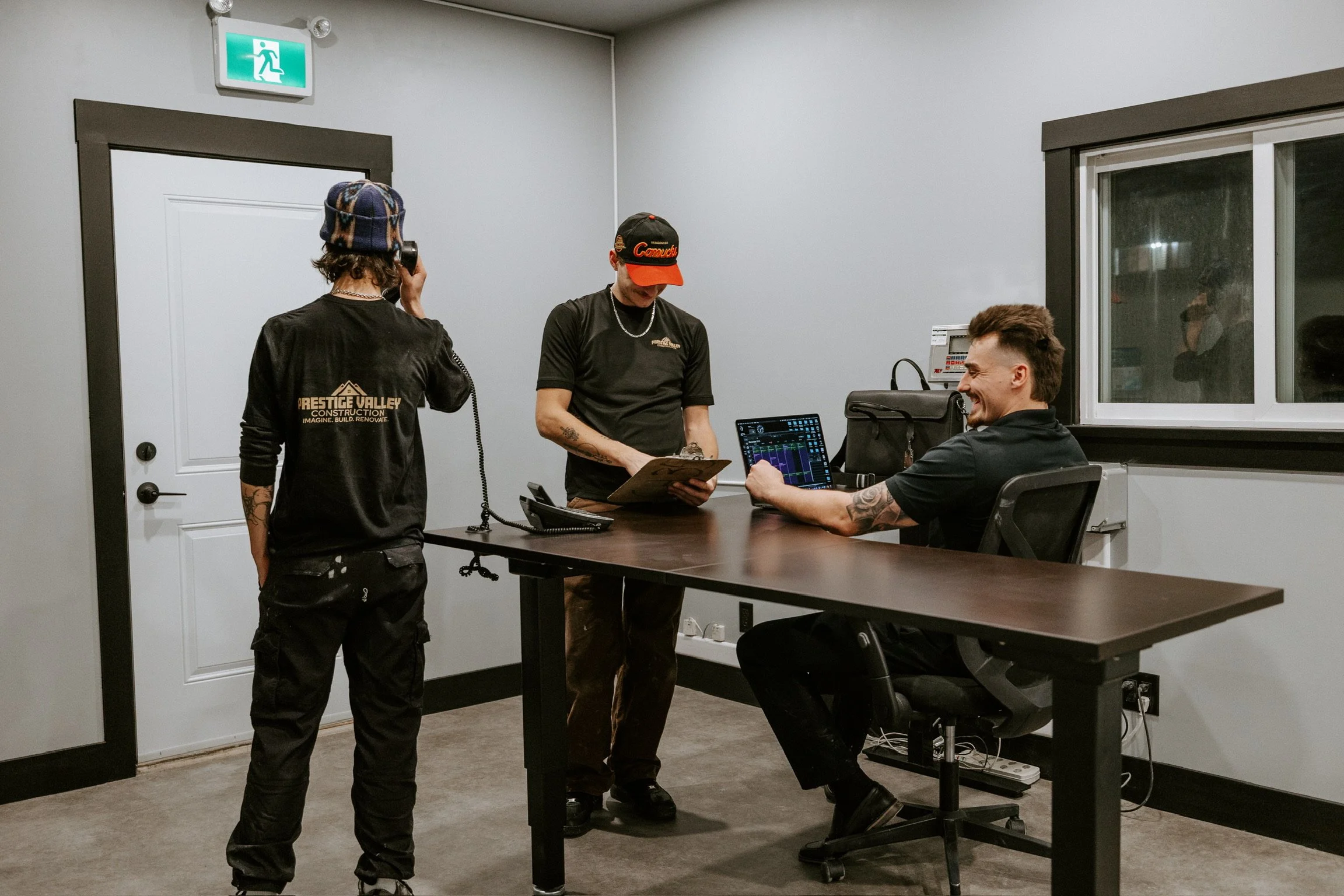 Three young men in a meeting room, one is seated at a desk with a laptop showing colorful graphs, another is standing with a clipboard, and the third is talking on a phone with a headset, wearing a black long-sleeve shirt with 'Prestige Valley Construction' on the back.