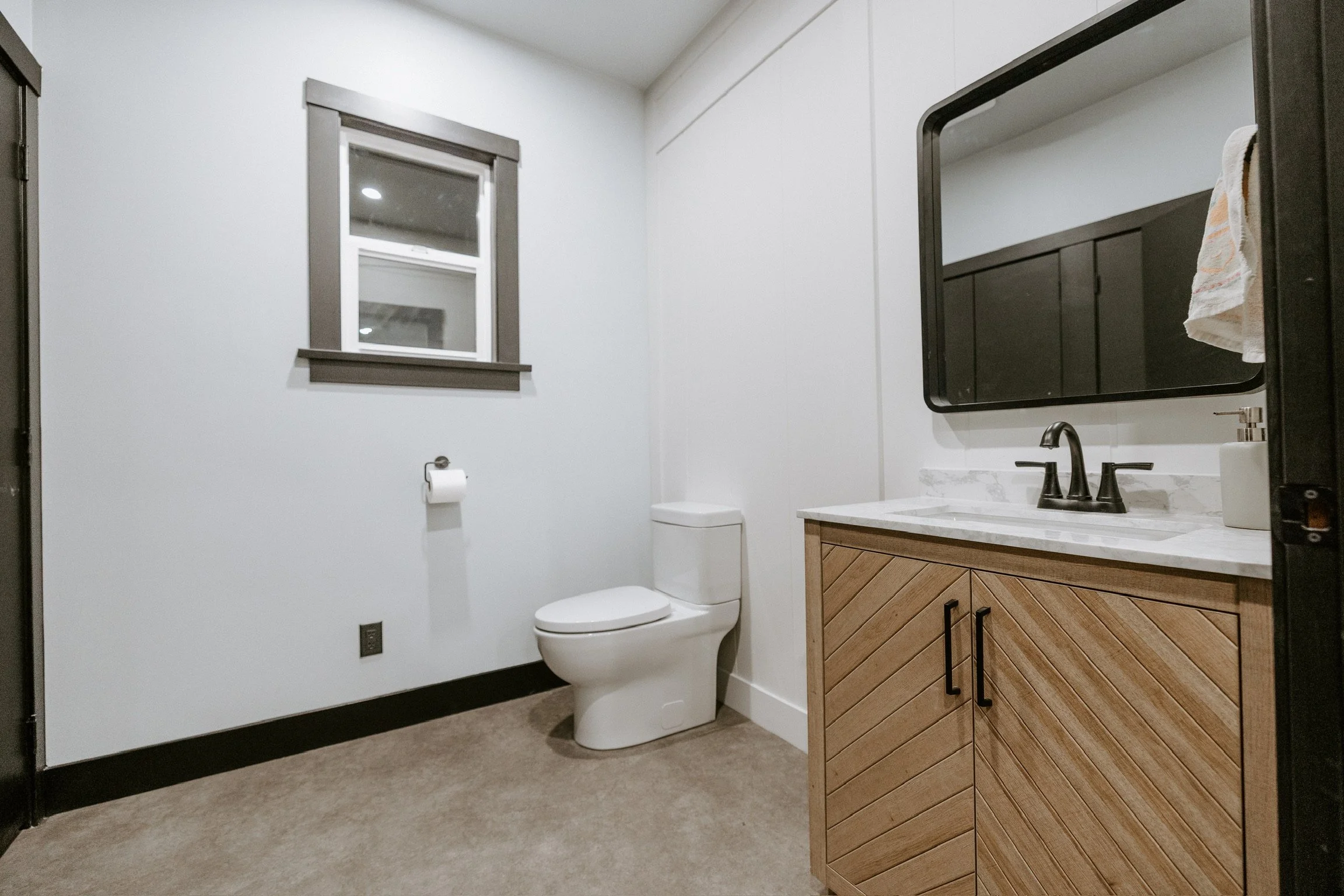Bathroom with white walls, a window with dark trim, a white toilet, a mirror above a wooden vanity with a marble countertop, and a towel hanging on the side of the mirror.