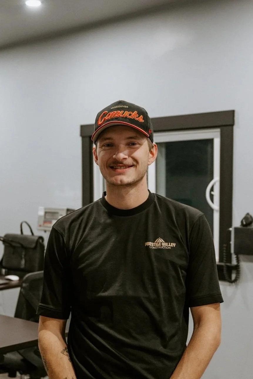 A young man smiling, wearing a black baseball cap with 'Vancouver Canucks' on it and a black T-shirt with the logo 'Prestige Valley Construction', standing indoors in front of a window and office equipment.