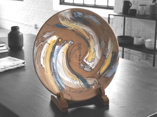 Decorative ceramic plate with abstract swirl patterns in blue, yellow, and white on a wooden display stand on a dark table.