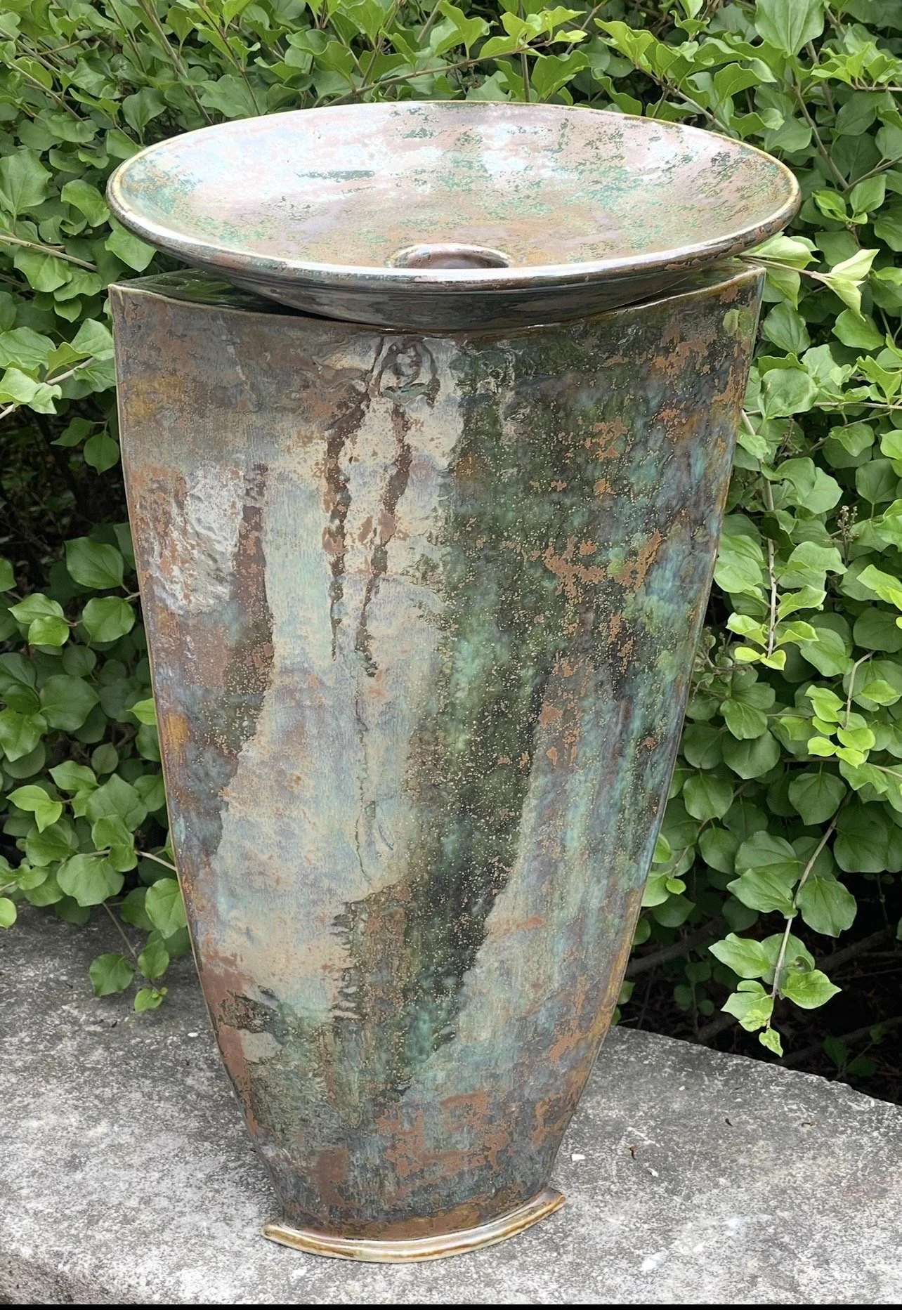 A tall, rusted metal sculpture with a wide, shallow bowl on top, positioned outdoors on a concrete surface with green foliage in the background.