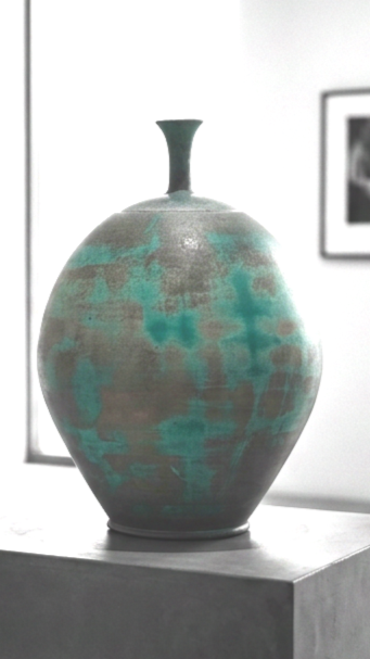 A ceramic vase with a narrow neck and rounded body, featuring a textured surface with teal and grayish-brown colors, placed on a pedestal in a gallery or museum setting.