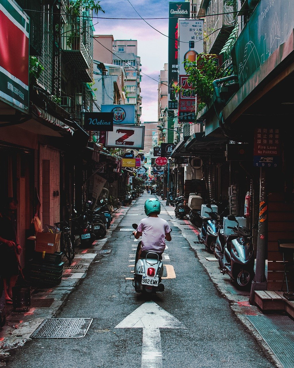 A person riding their white scooter down a narrow street in Taiwan lined with other scooters and large shop signs during a Taiwan Cultural Experience with Tricia Pomering..