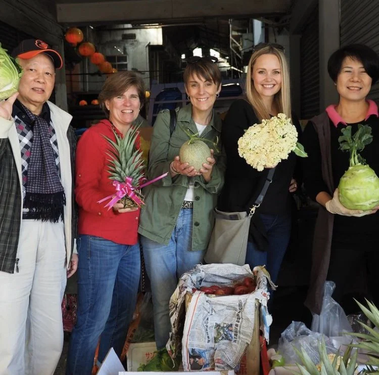 Tricia Pomering with two European friends standing with two Taiwanese individuals at their vegetable stand in Taiwan during a Cultural Immersion Experience hosted by Tricia. Each person is holding a different vegetable and smiling.