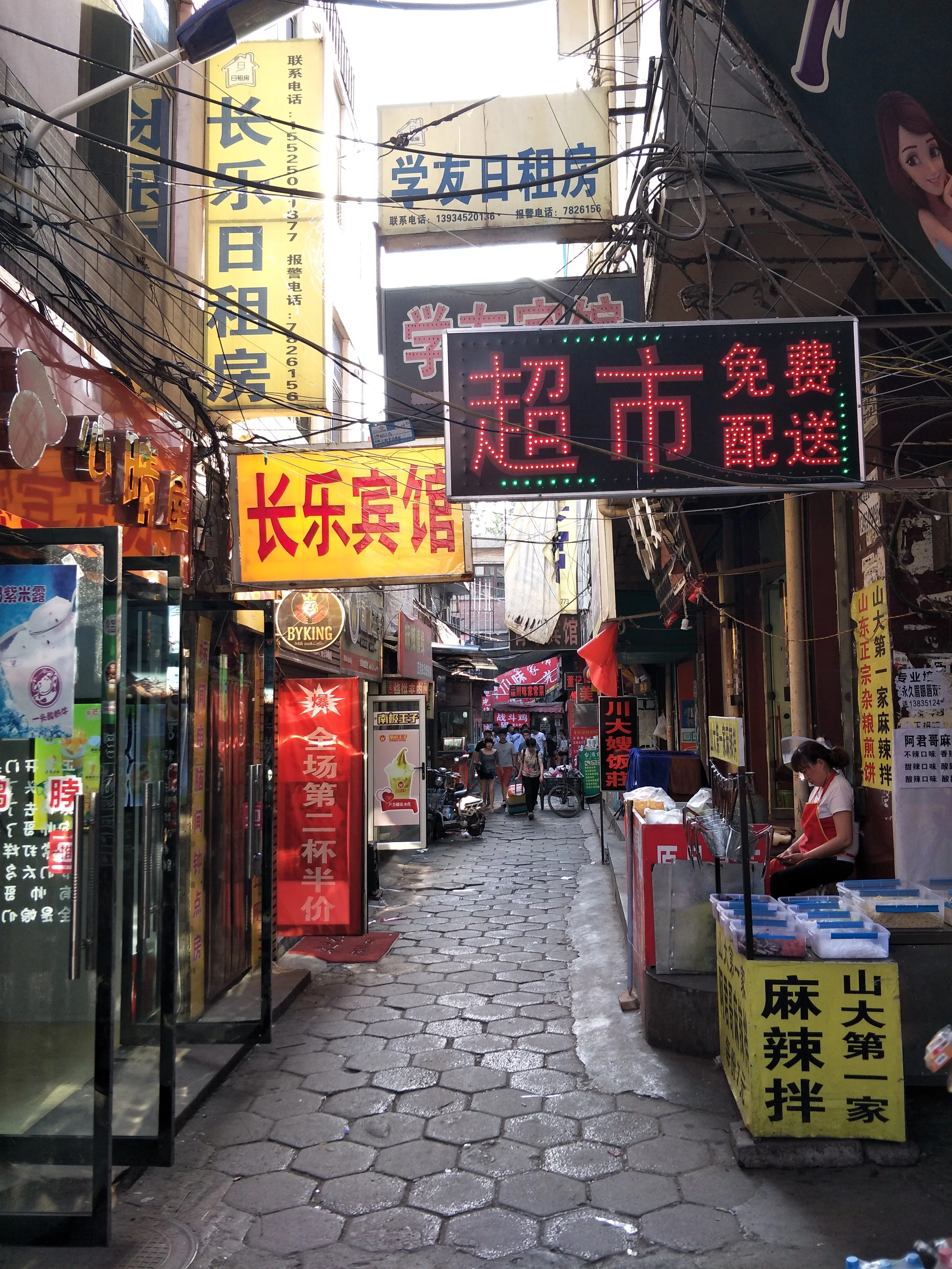 Day market with food stalls, large bright yellow and red signs, and a woman waiting to sell items in Taipei during a Tricia Pomering culture retreat.