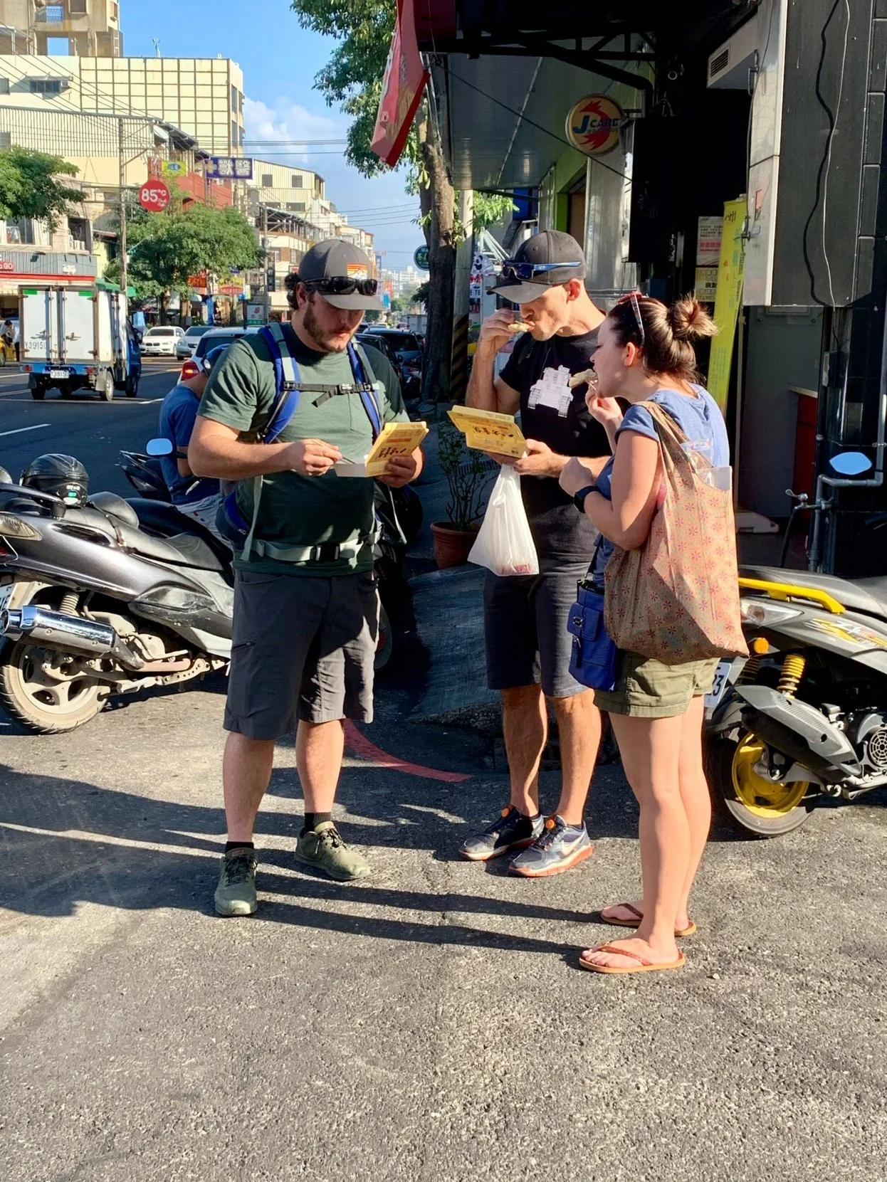 A group of individuals stand on a sidewalk in Taiwan tasting local foods during a Cultural Experience with Tricia Pomering.