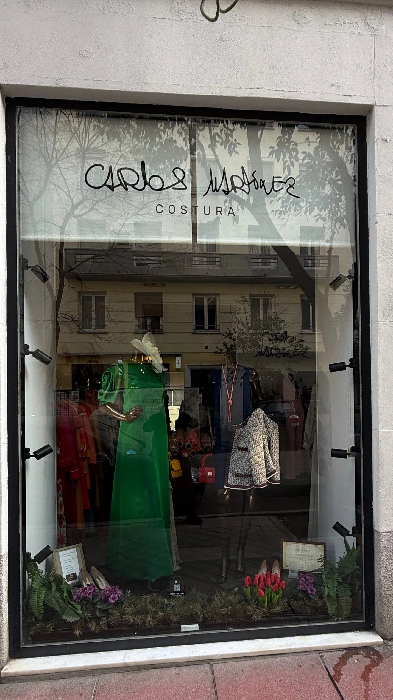 Vitrina de tienda de ropa con el nombre 'Carlos Muñoz Costura'. Dentro se ven prendas de ropa en exhibición, incluyendo un vestido verde, un abrigo con detalles en negro y blanco, y accesorios. En la parte inferior, hay plantas y flores decorativas.