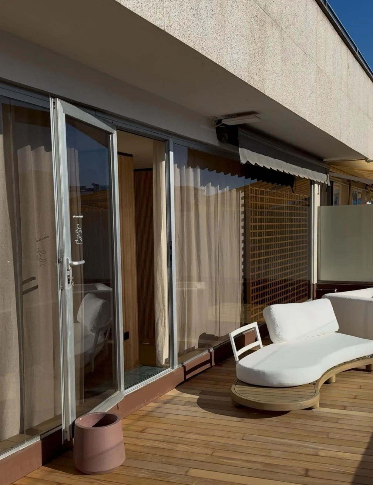 Terraza de un edificio con sillón curvo de madera y cojines blancos, cerca de ventanas de vidrio con cortinas beige, y una maceta rosada.