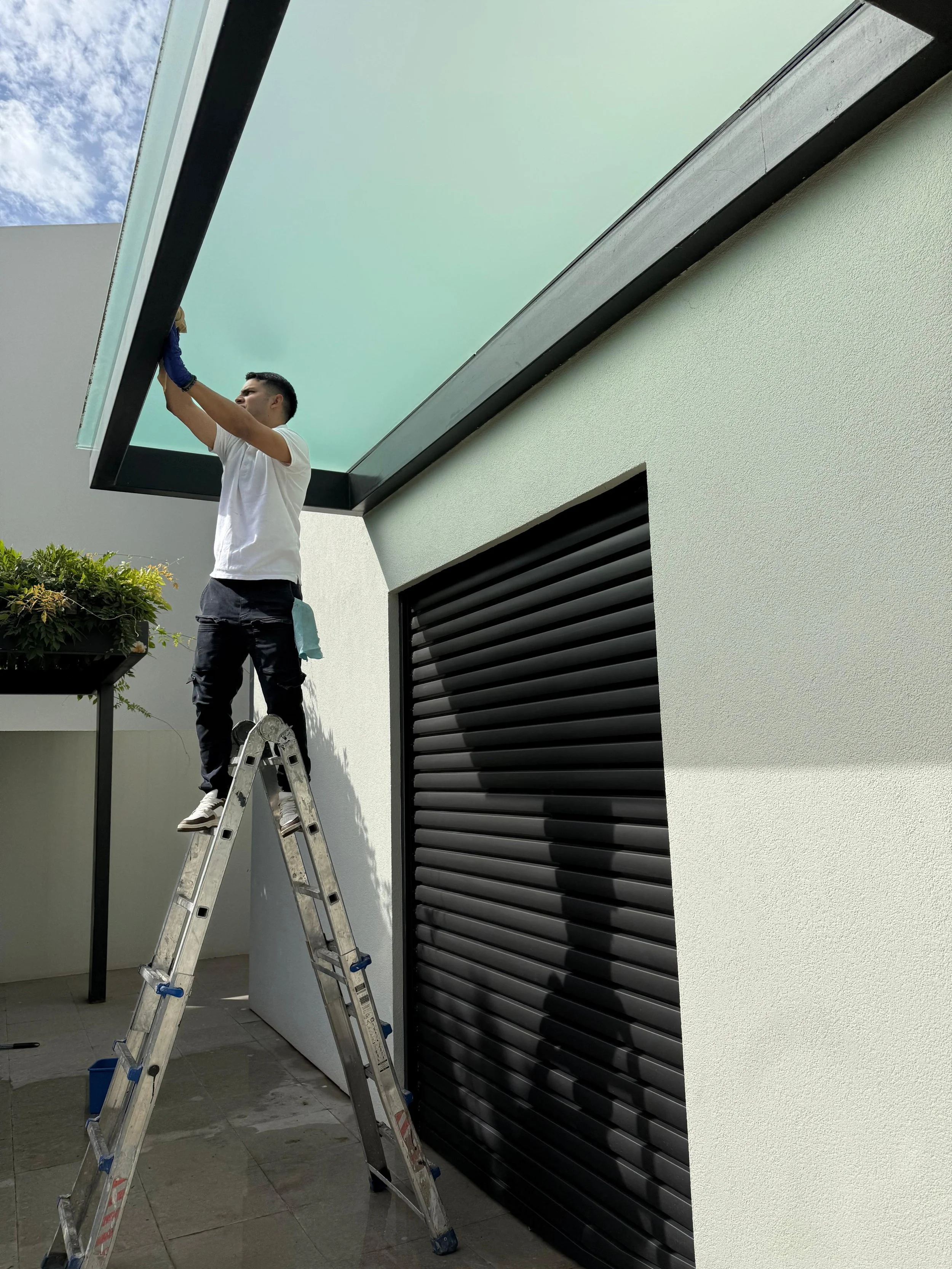 Hombre en una escalera limpiando o reparando el marco de un ventanal grande en una casa moderna.