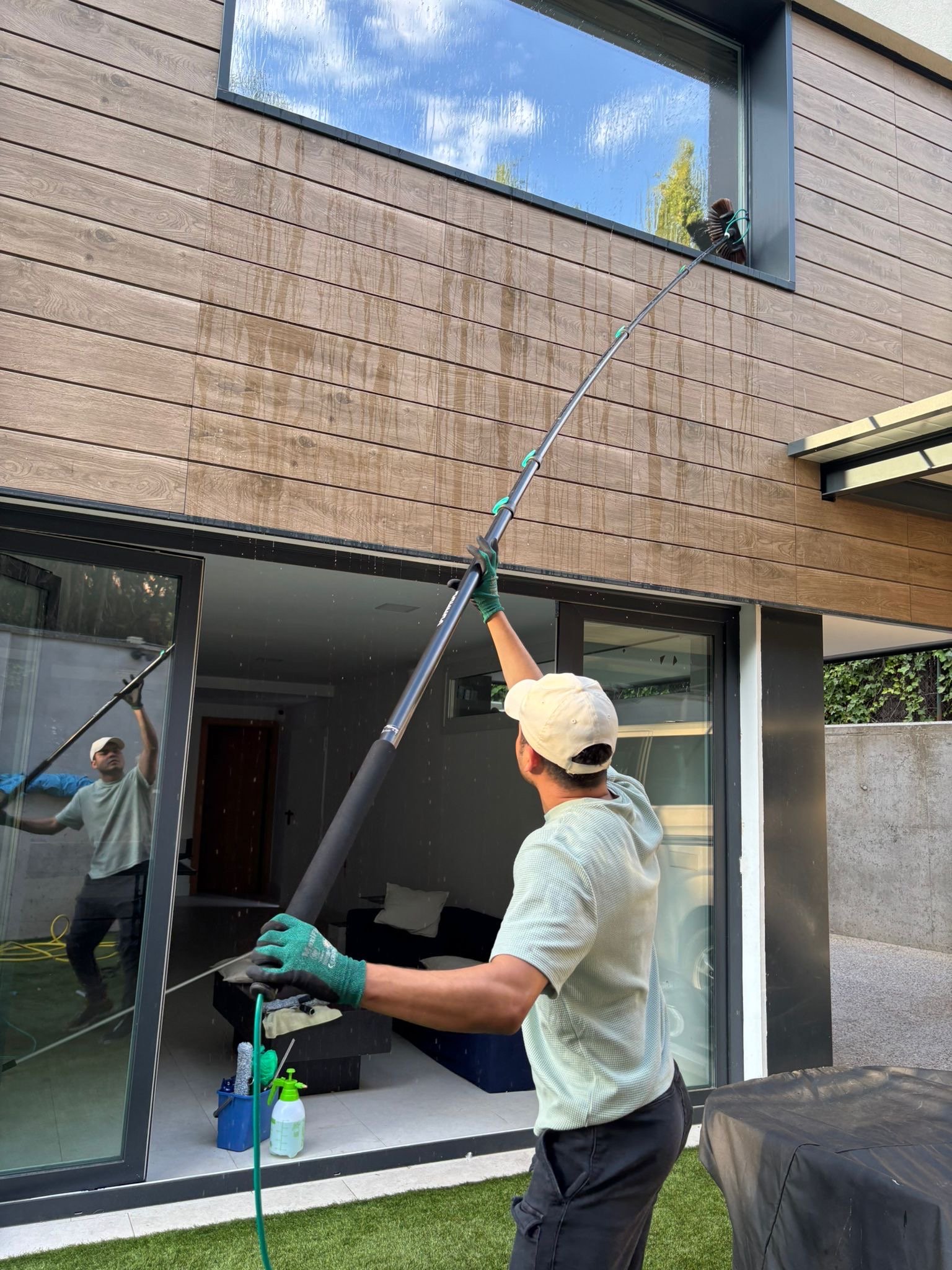 Persona limpiando un ventanal grande con un utensilio largo. Se observa reflejo del cielo y árboles en el cristal. La escena ocurre en una casa moderna con exterior de madera y vidrio, con otros utensilios de limpieza en el entorno.