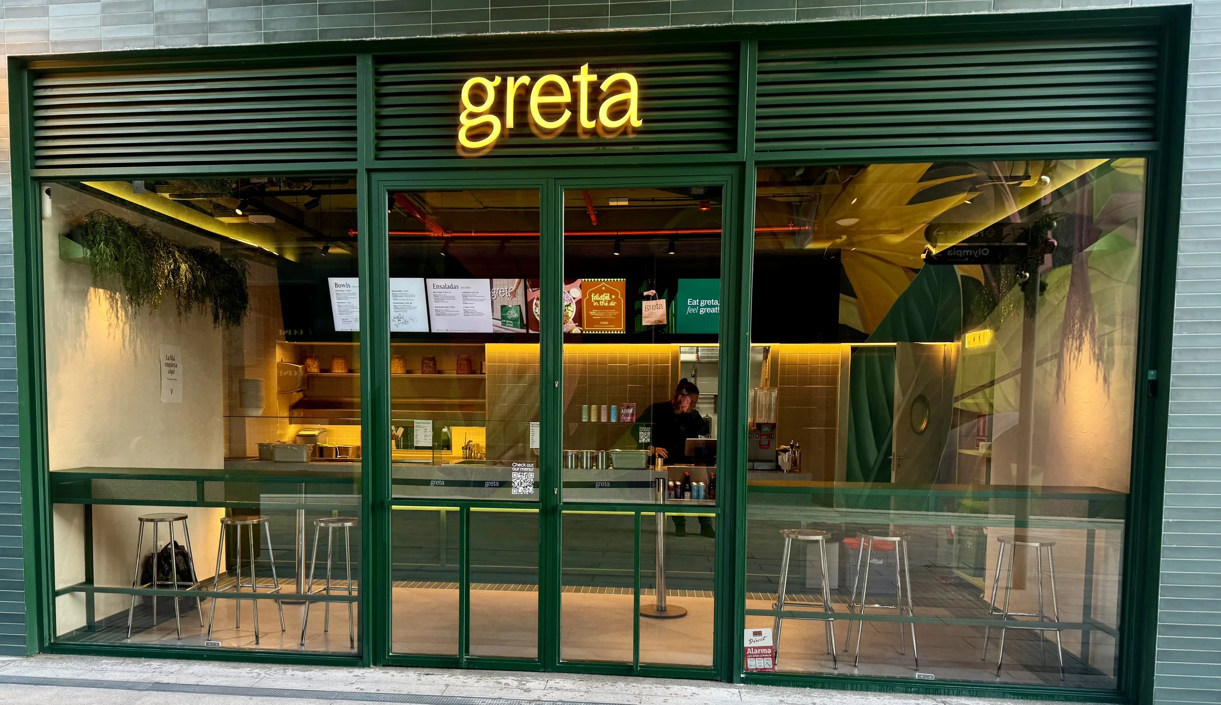 Fachada de un restaurante llamado Greta con ventanales grandes, interior visible con barra y mesas, y decoraciones con plantas y colores verdes y amarillos.