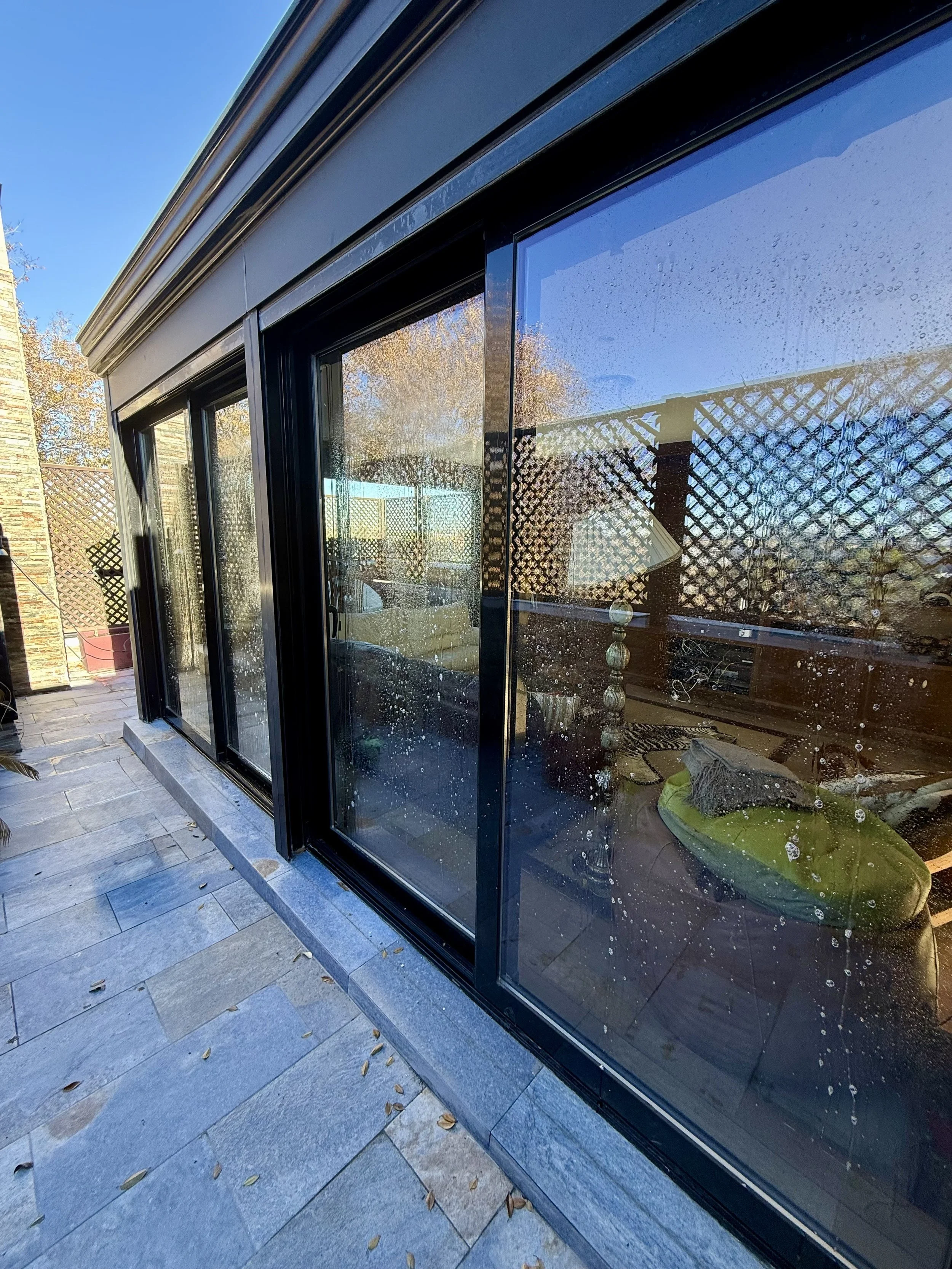 Ventanas de vidrio con gotas de agua impactando y reflejando el entorno, incluyendo decoración interior y plantas en un espacio exterior adoquinado.