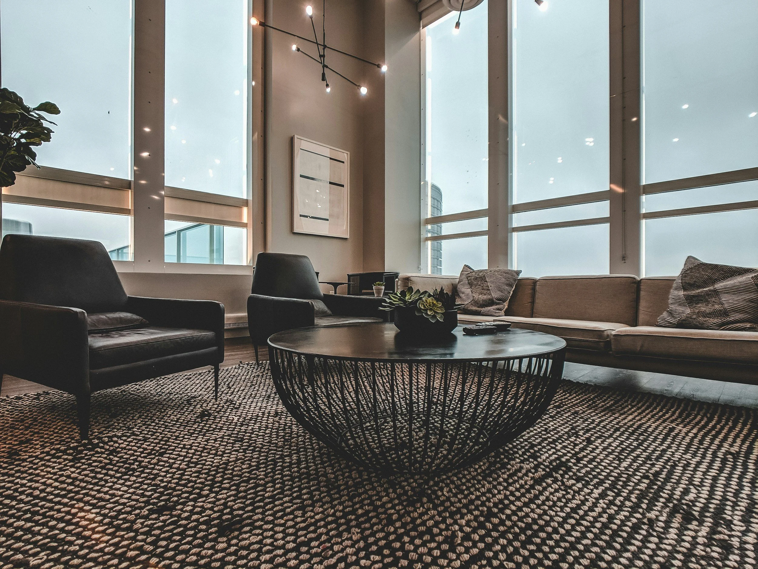 Modern living room with large floor-to-ceiling windows, black leather armchairs, a beige sofa with cushions, a round black wire coffee table with a plant, and a patterned rug.