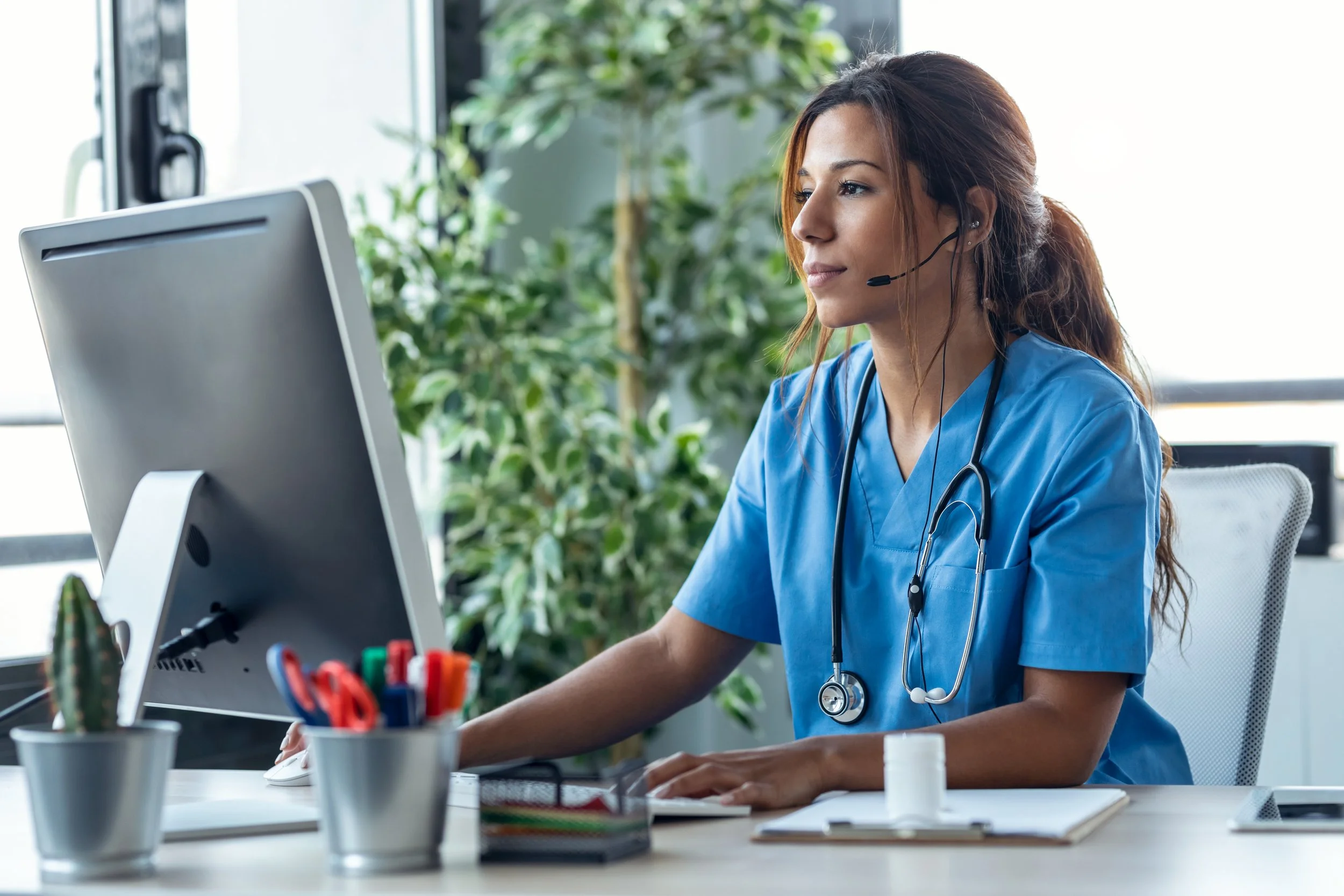 Woman nurse sitting at desk on a computer wearing a headset.