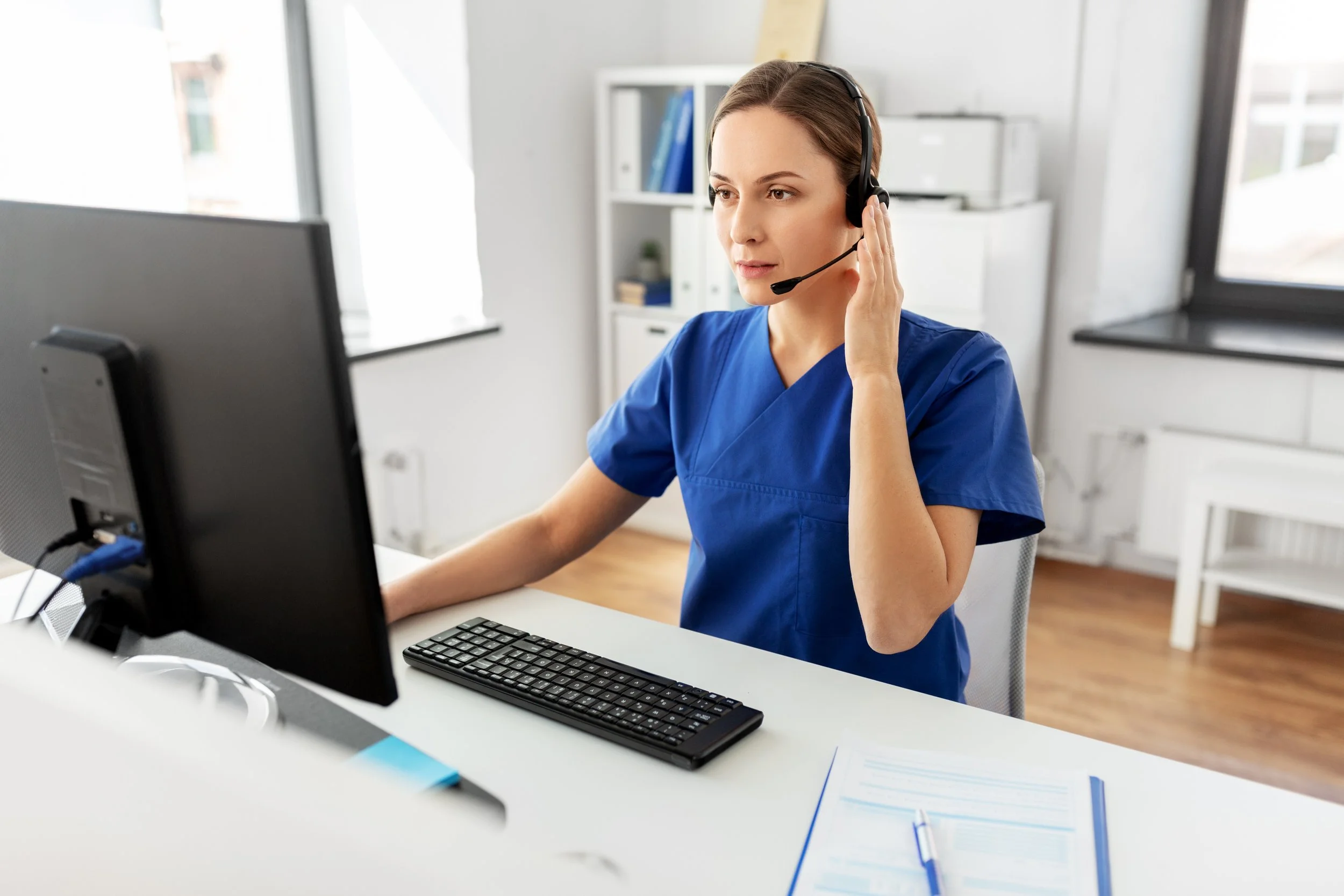 Woman nurse wearing a headset at a desk working on a computer.