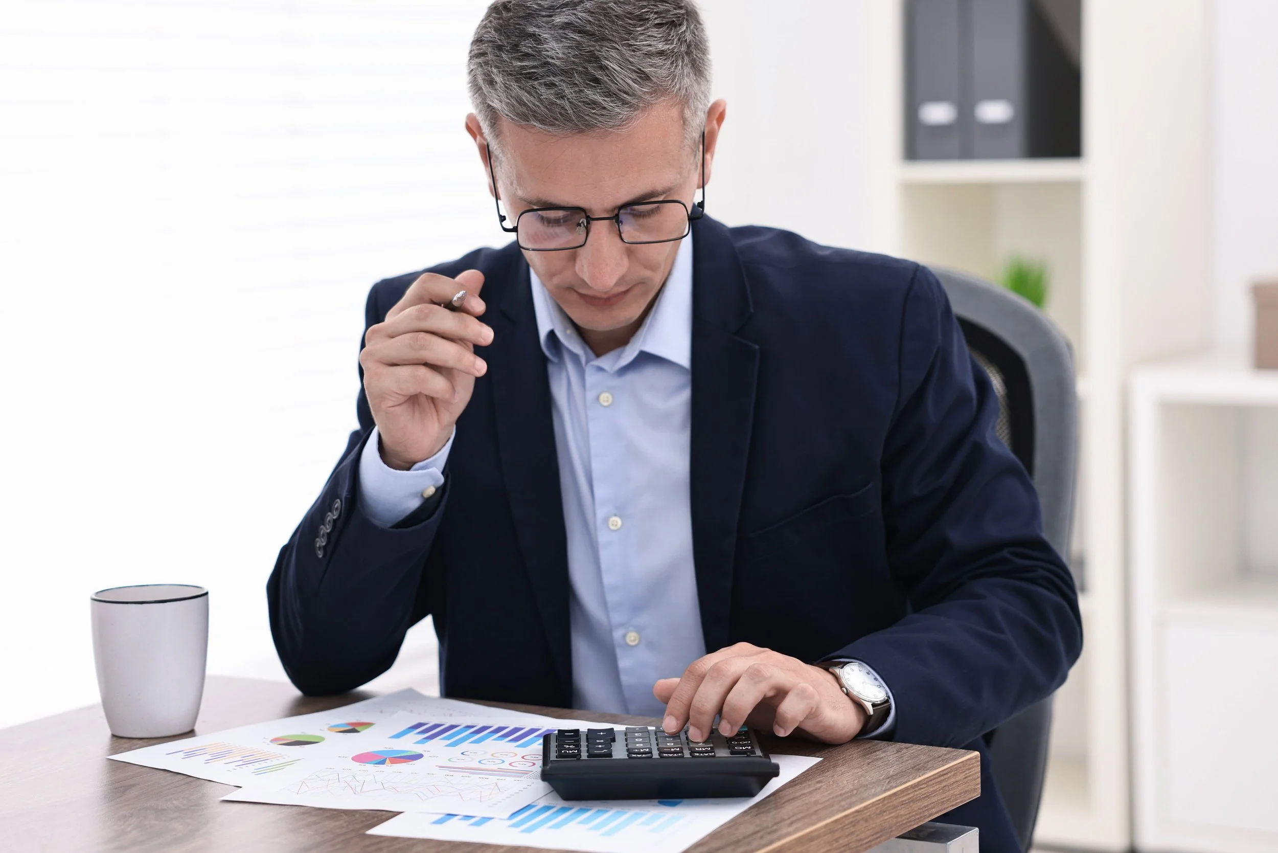 Businessman sitting at a desk typing on a calculator with papers underneath displaying financial graphics.