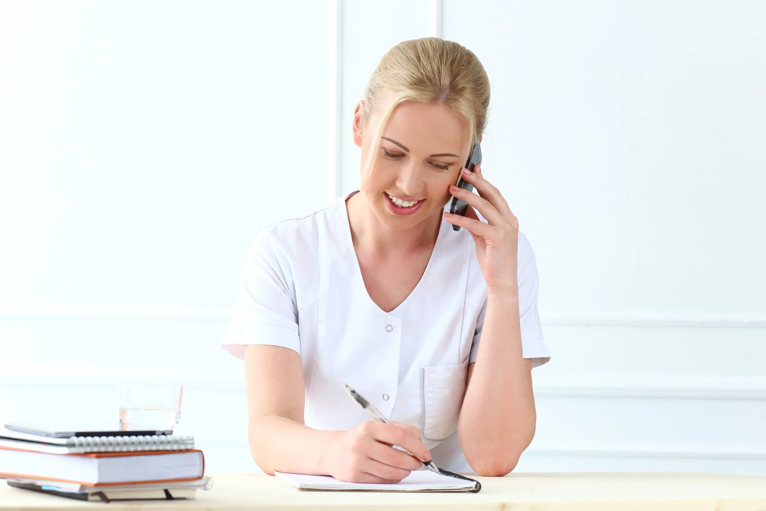 Women nurse talking on her cellphone at a desk while writing with a pen in a notebook.