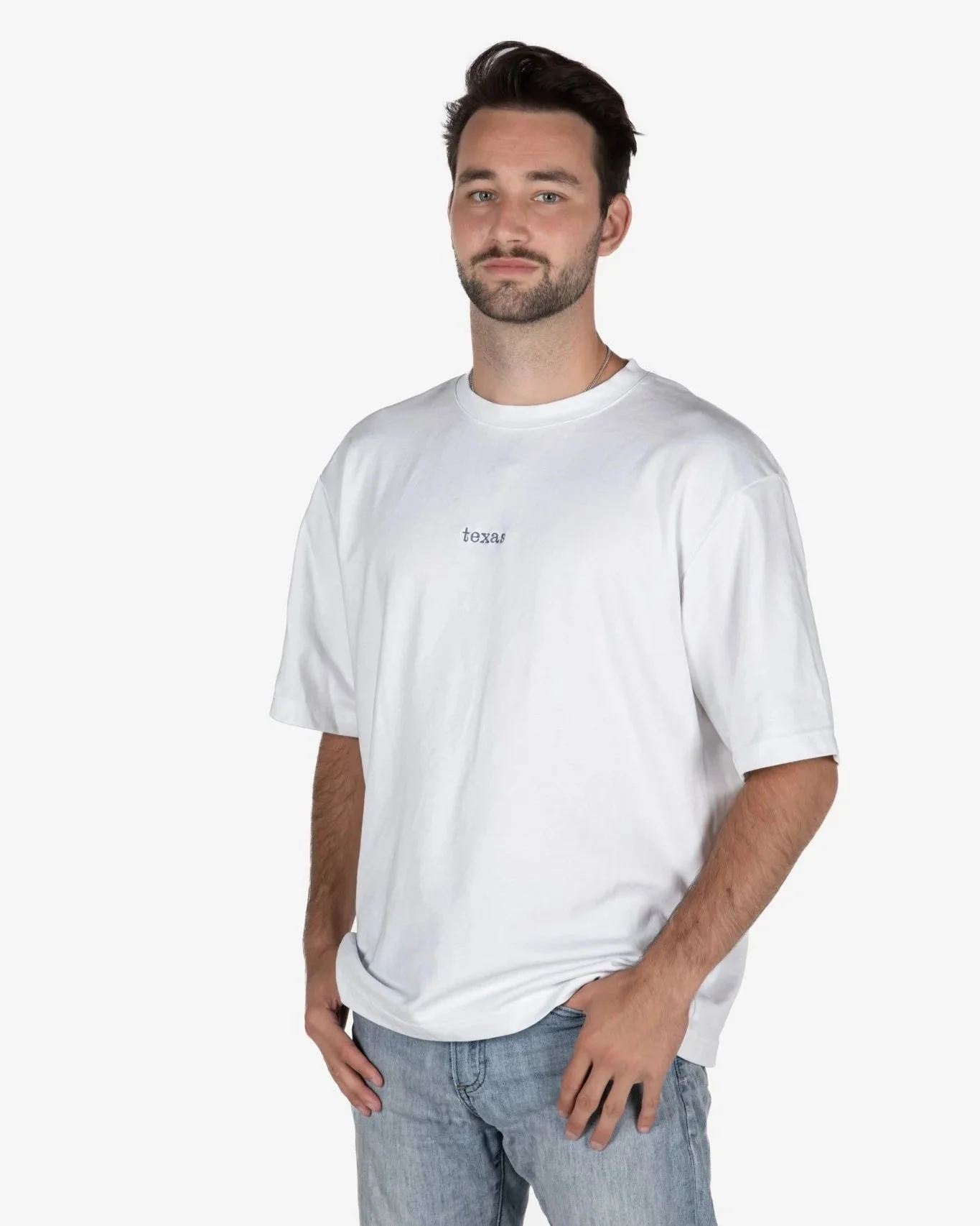 Man with short dark hair and beard wearing a white singular clothing T-shirt embroidered with the word "texas", standing against a plain white background.