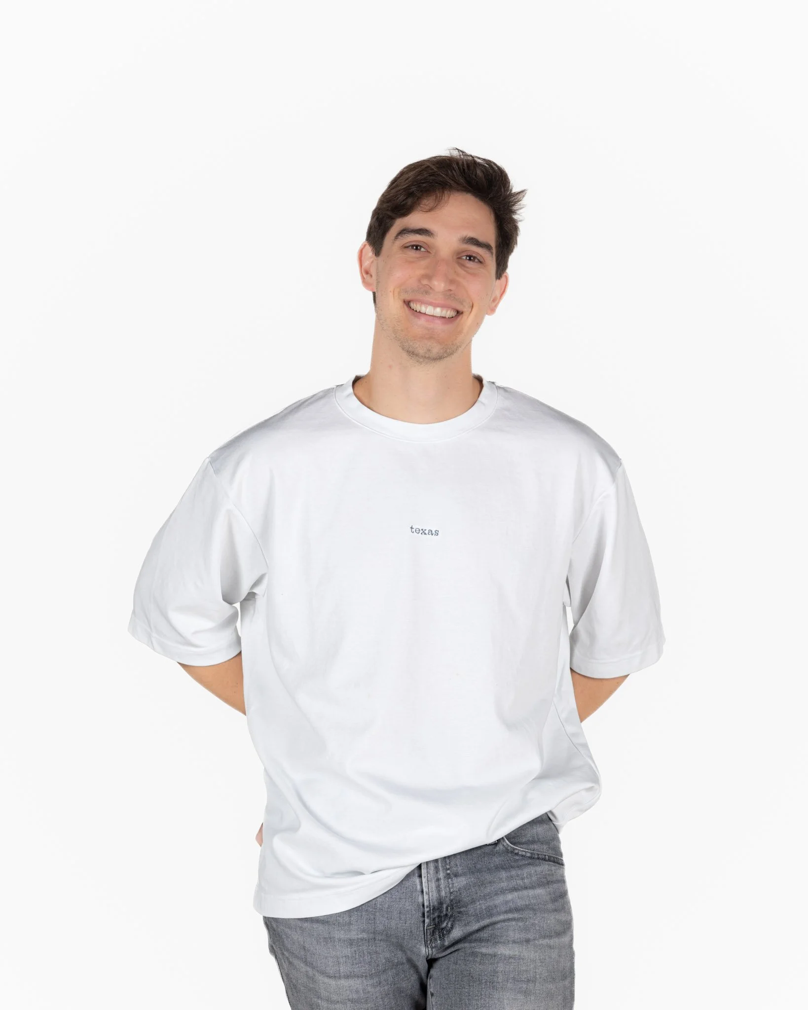 A young man with dark hair, smiling and wearing a white singular clothing T-shirt with the word "texas", standing against a plain white background.