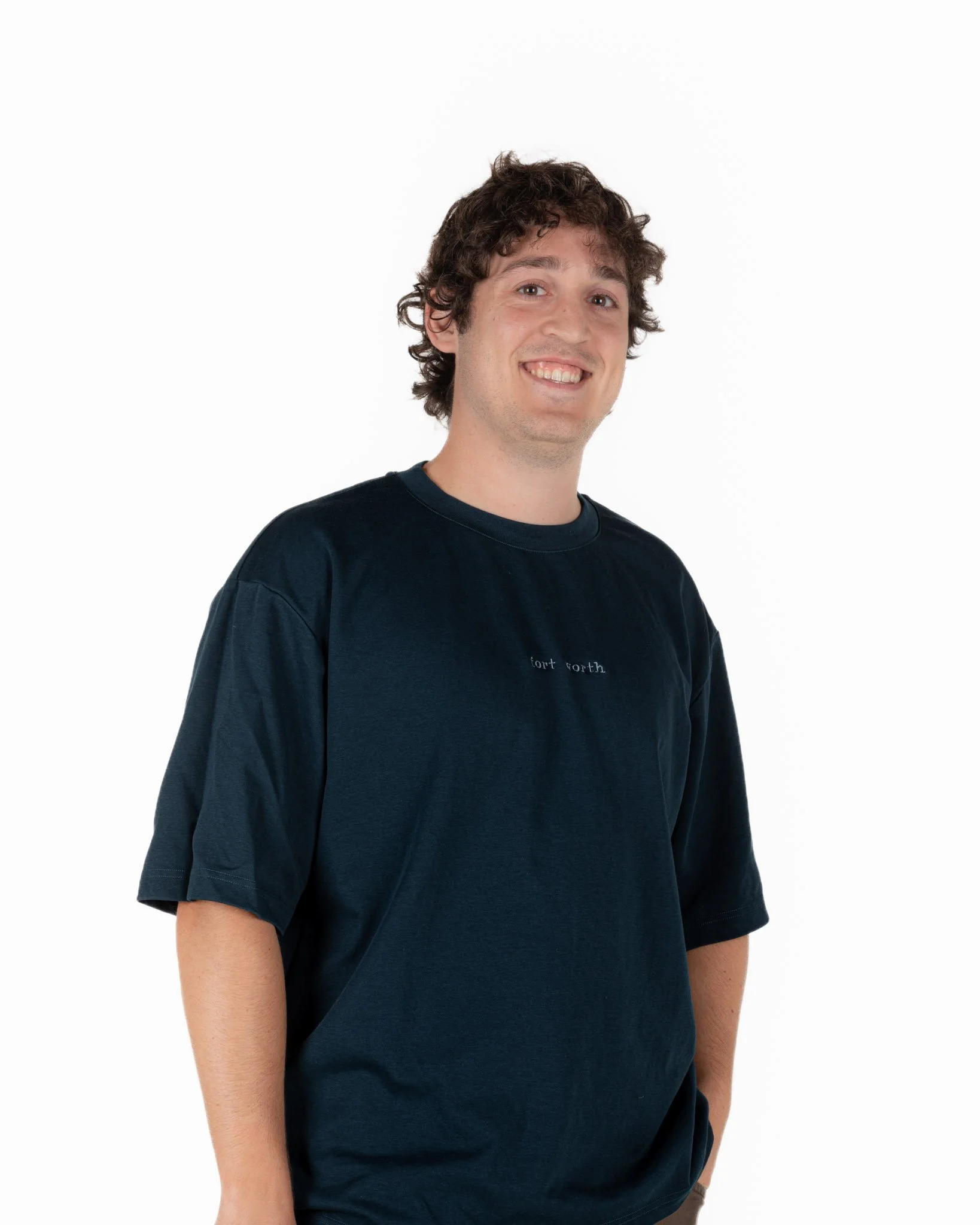 A man with curly brown hair wearing a blue singular clothing T-shirt embroidered with the word "fort worth", smiling, and standing against a white background.
