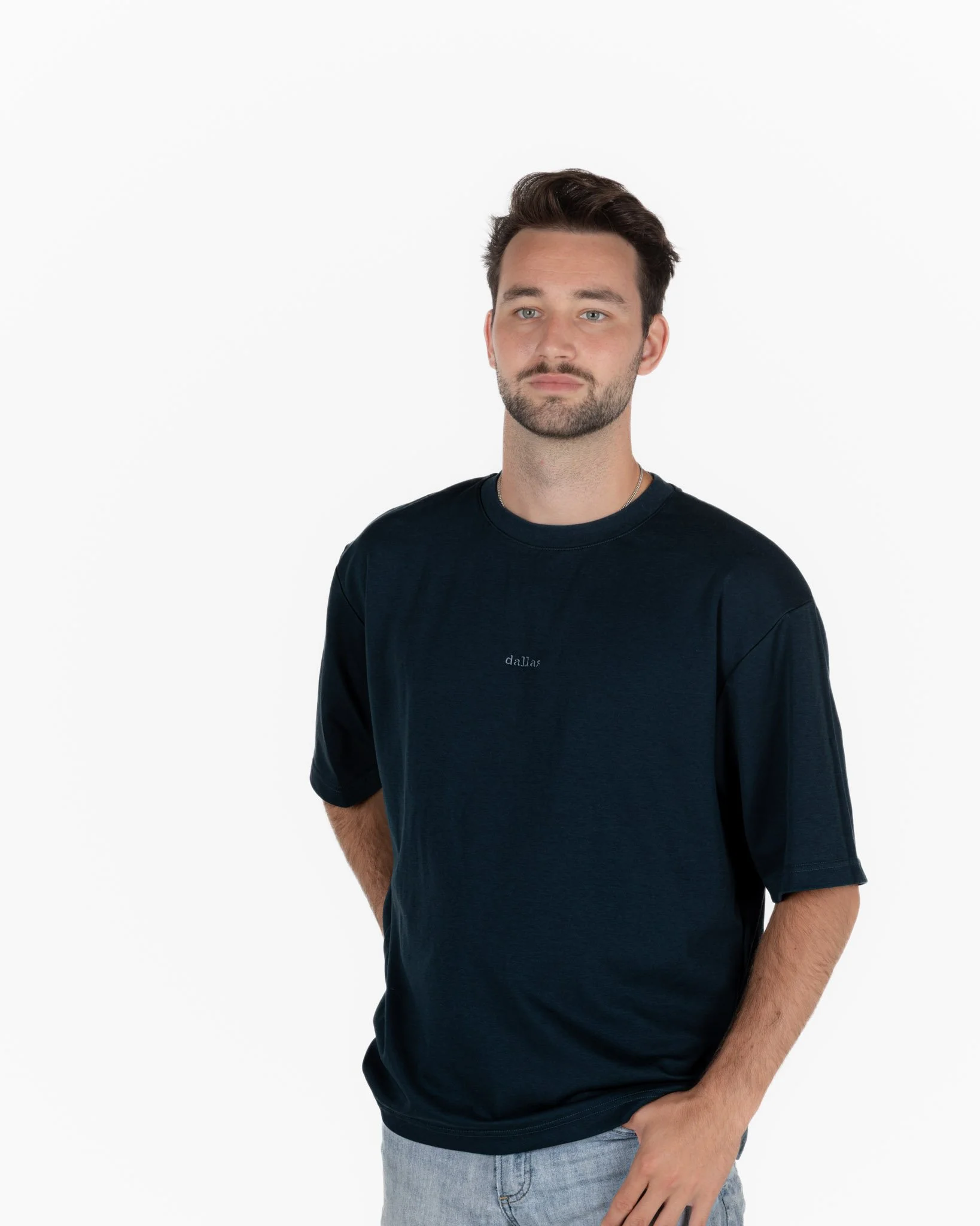 A young man with dark hair and a beard, wearing a blue singular clothing T-shirt embroidered with the word "dallas", standing against a plain white background.