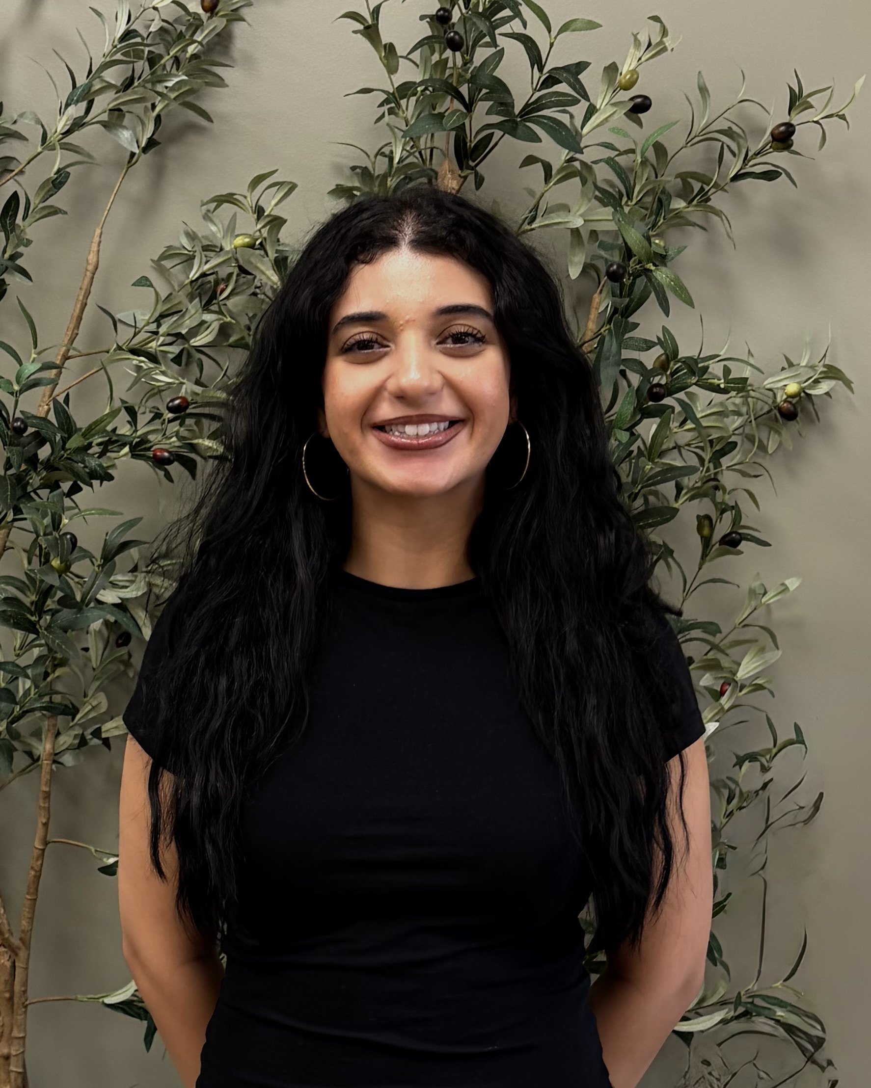 A woman with long black hair and hoop earrings standing in front of an olive tree with green and black olives, wearing a black shirt.