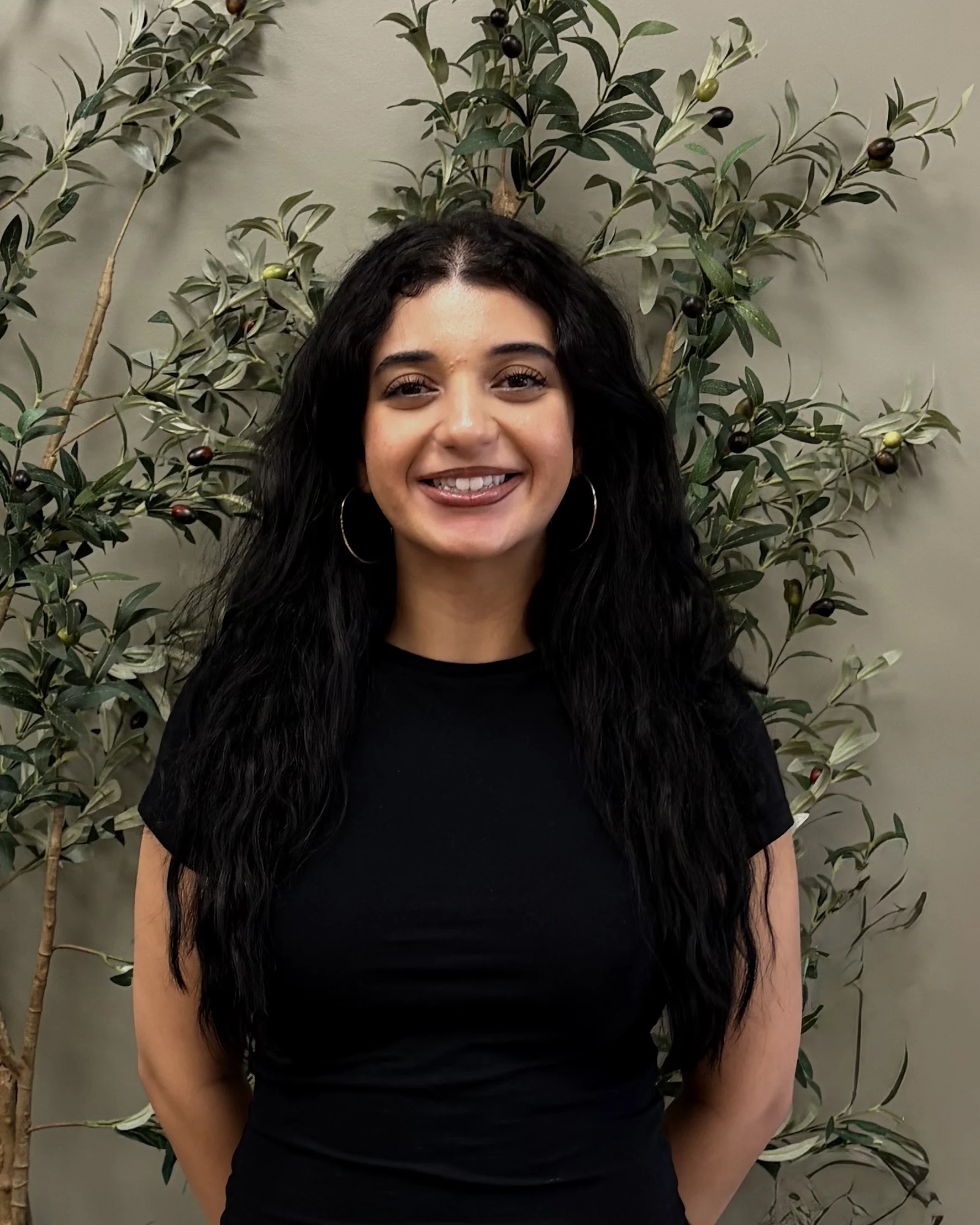 Headshot of a woman with long black hair, smiling, wearing a black shirt and hoop earrings, standing in front of a leafy green plant with small dark and green berries.