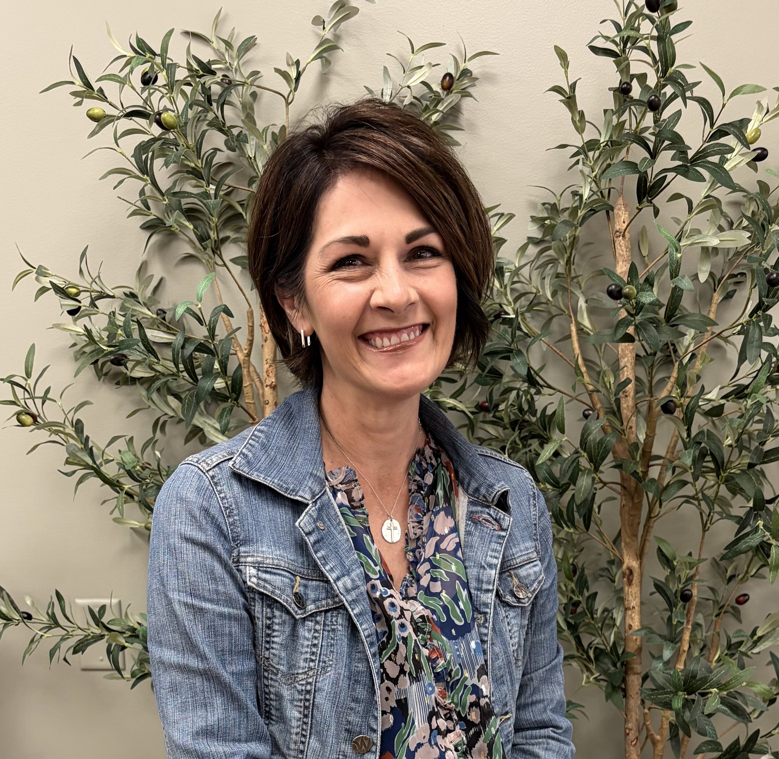 A woman with brown hair smiling, wearing a denim jacket and floral shirt, standing in front of a decorative olive tree with green and black olives against a beige wall.