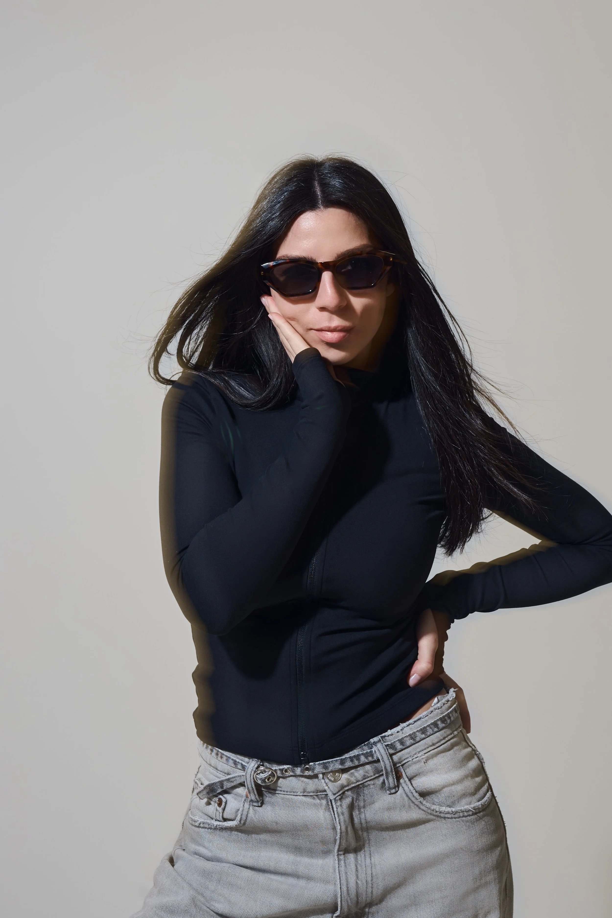 Nahes Portrait vor hellem Hintergrund. Person im schwarzen Zip-Up mit Sonnenbrille stützt das Gesicht auf die Hand.