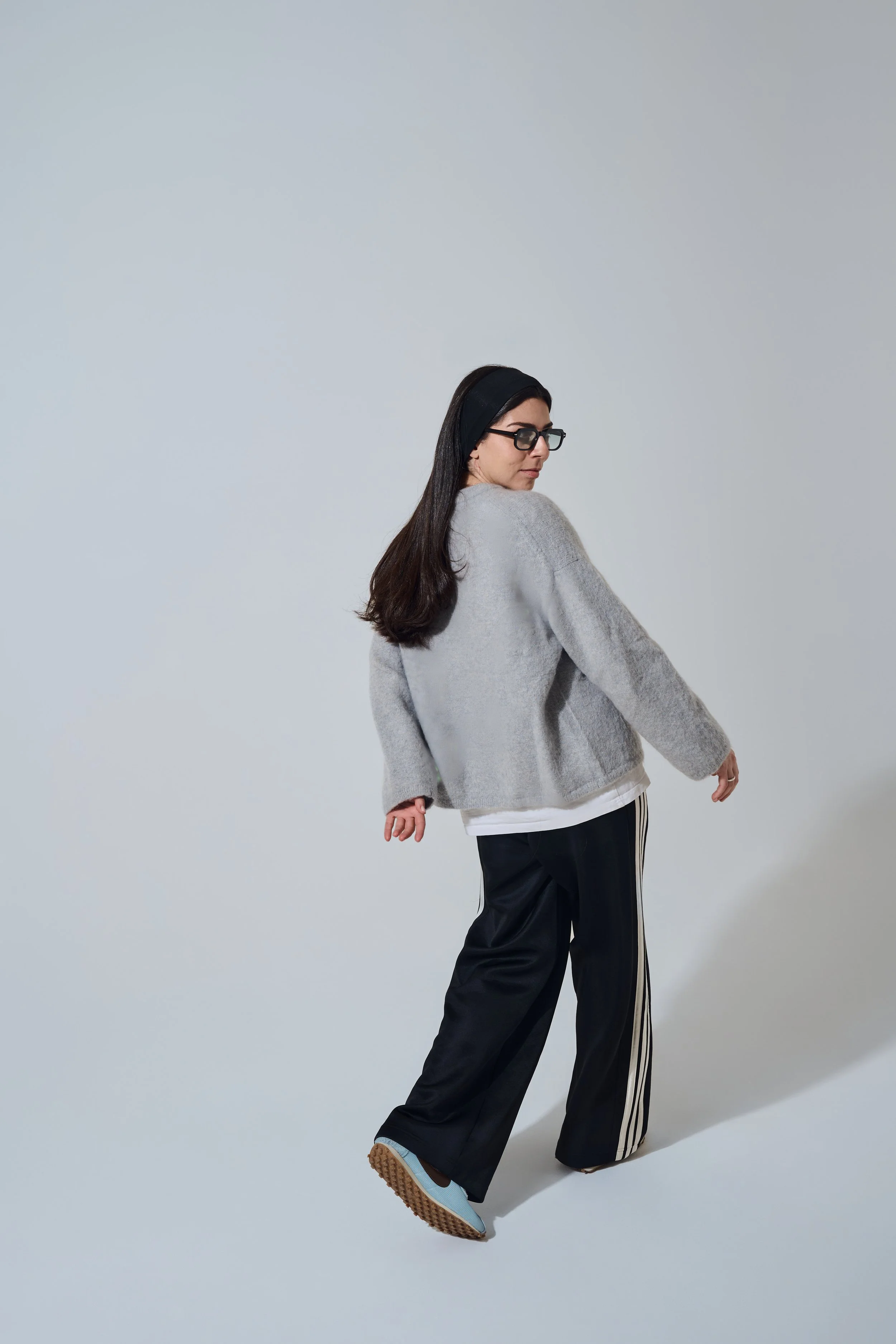 Rückansicht beim Gehen im Studio. Person im grauen Pullover und schwarzer Streifenhose blickt über die Schulter zurück.