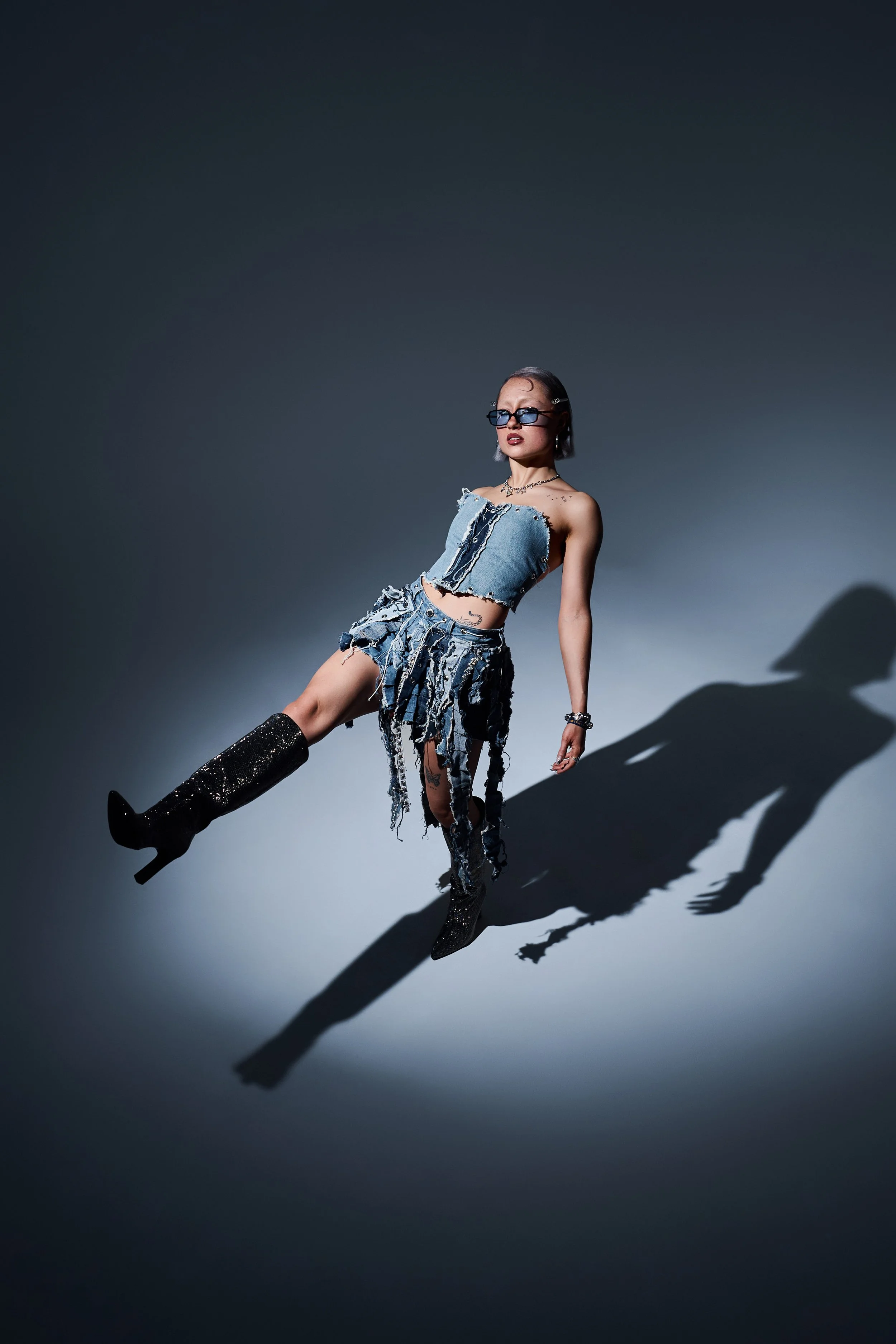 Dynamische Pose im Studio. Person kickt im Denim-Look und Glitzerstiefeln vor grauem Hintergrund mit hartem Schlagschatten.
