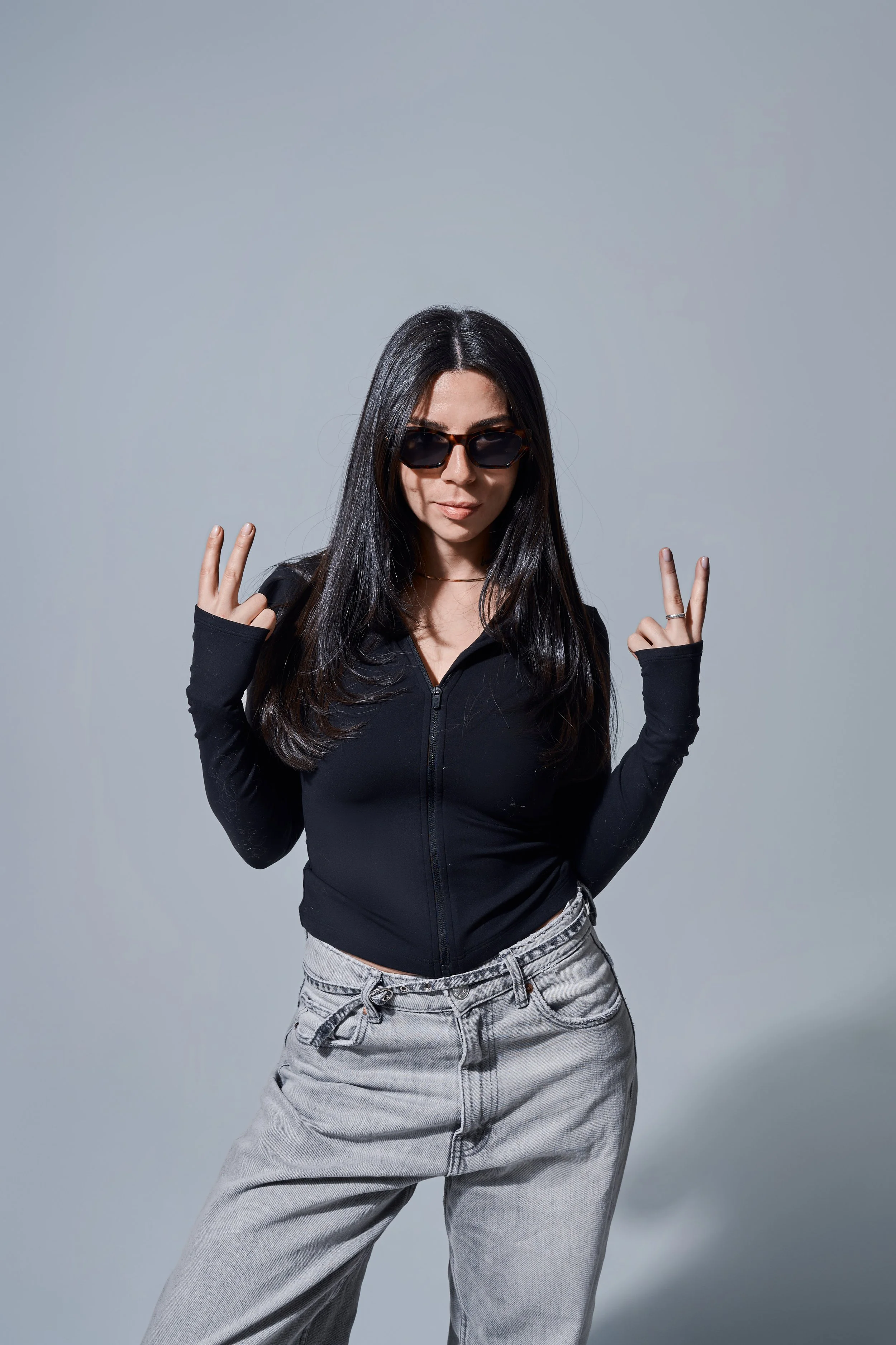 Portrait vor grauem Hintergrund. Person im schwarzen Oberteil und hellen Jeans zeigt mit beiden Händen das Victory-Zeichen.