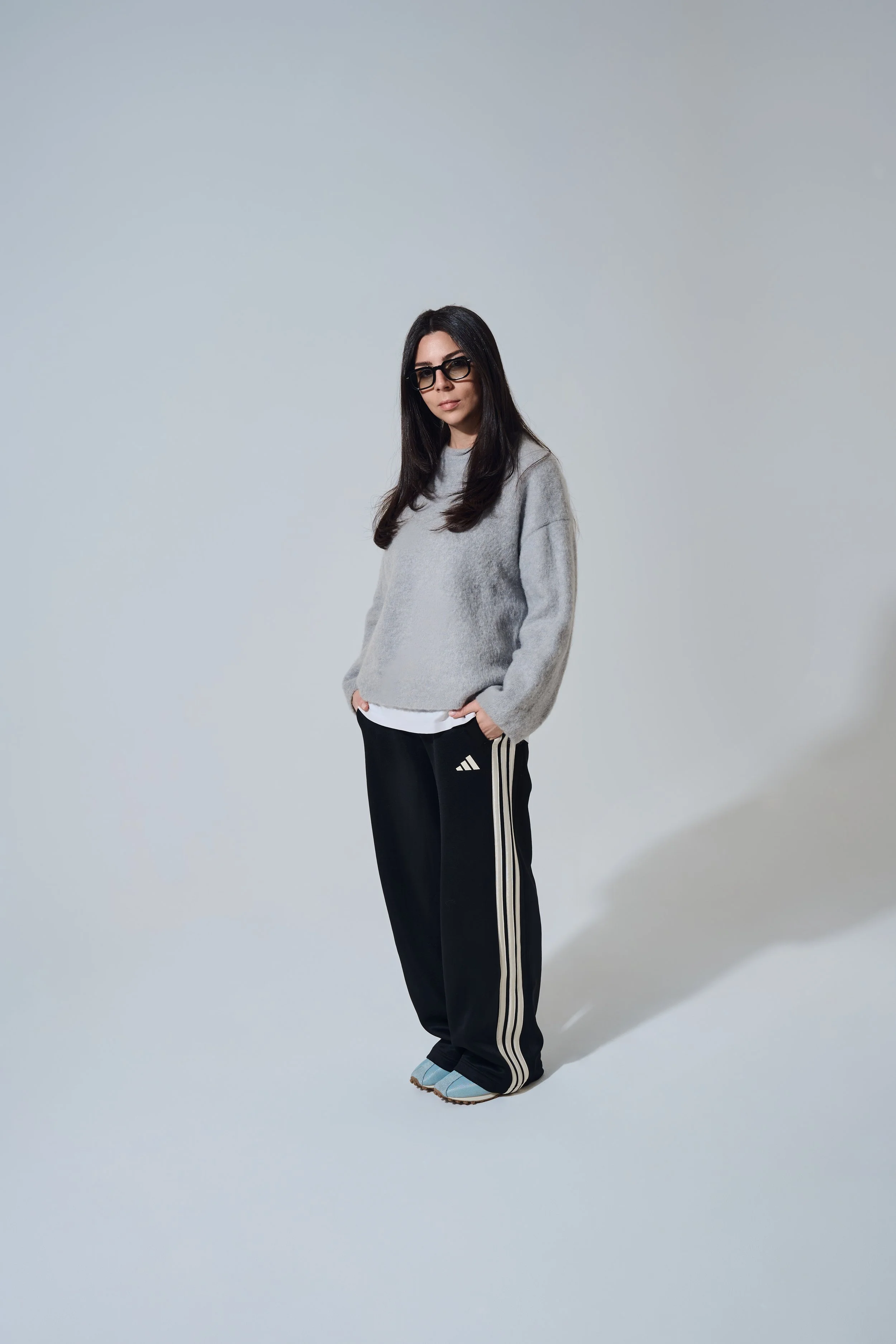 Ganzkörper-Portrait vor weißem Hintergrund. Person im grauen Strickpullover, schwarzer Trainingshose und Brille.