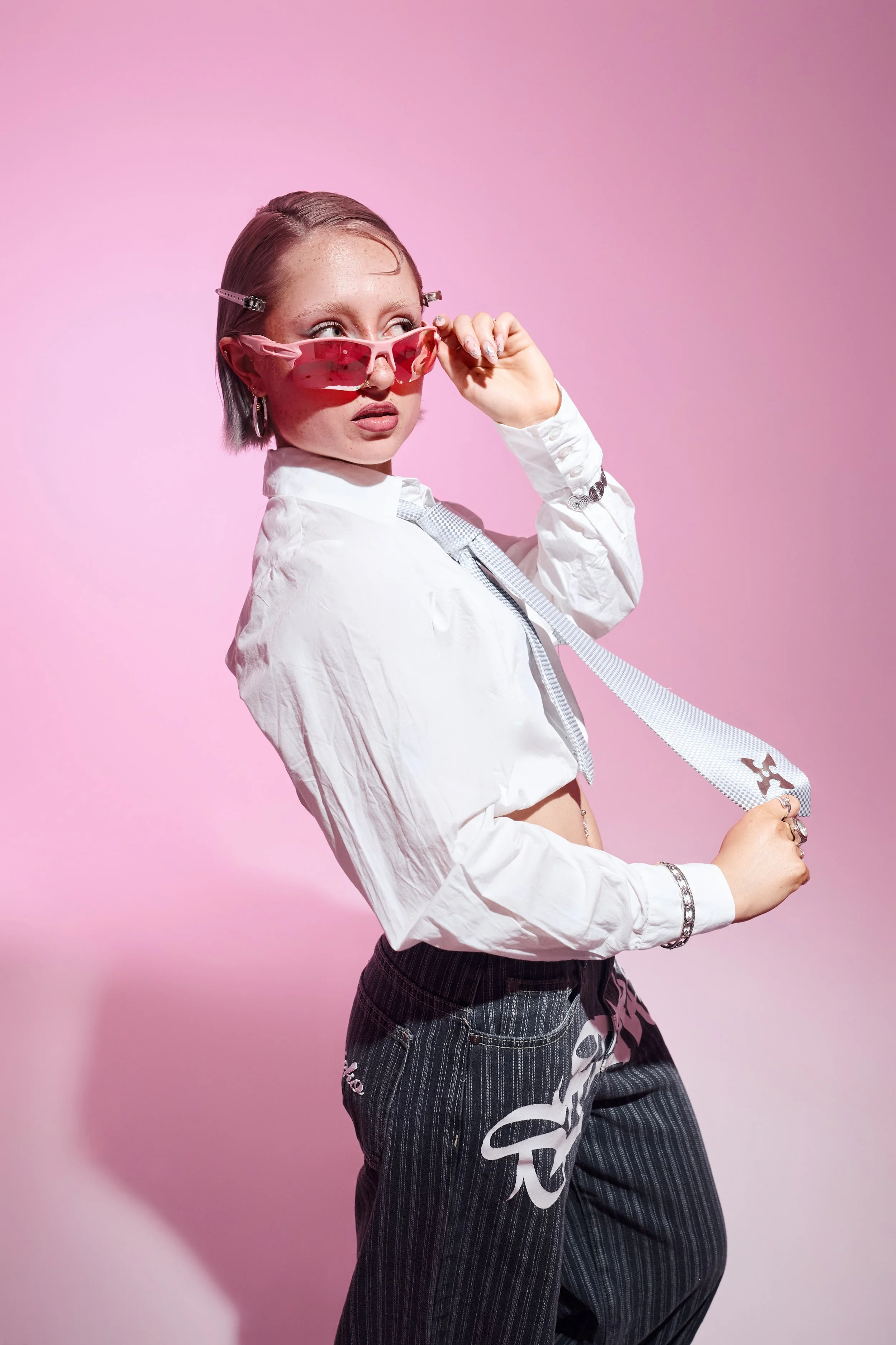 Fashion-Portrait im Studio vor pinkem Hintergrund. Person in weißem Kurzhemd und Krawatte hält eine rosa Sonnenbrille.
