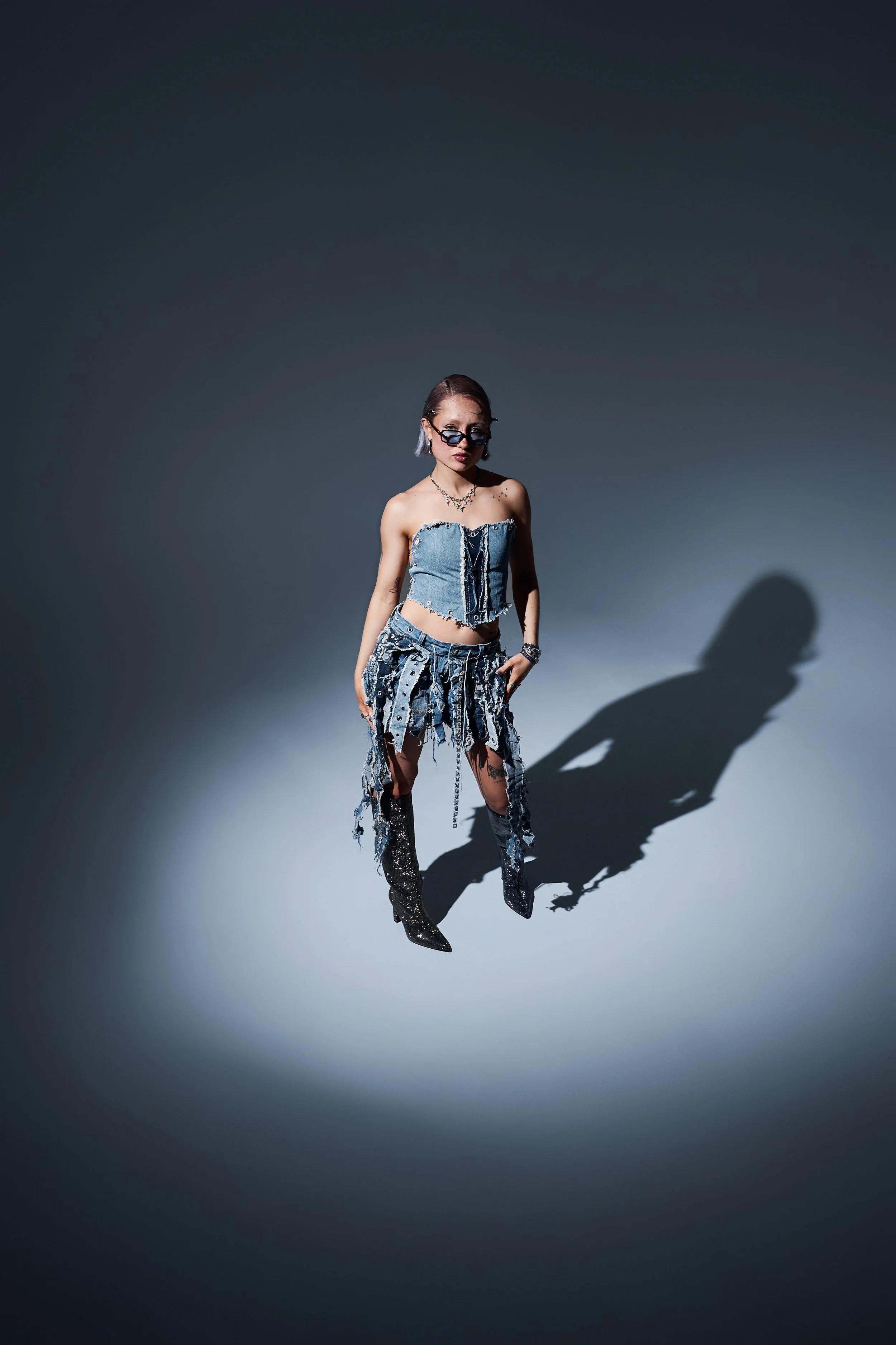 Ganzkörper-Fashion-Shot. Person im Denim-Outfit und dunklen Stiefeln steht in einem Lichtkegel auf grauem Studioboden.