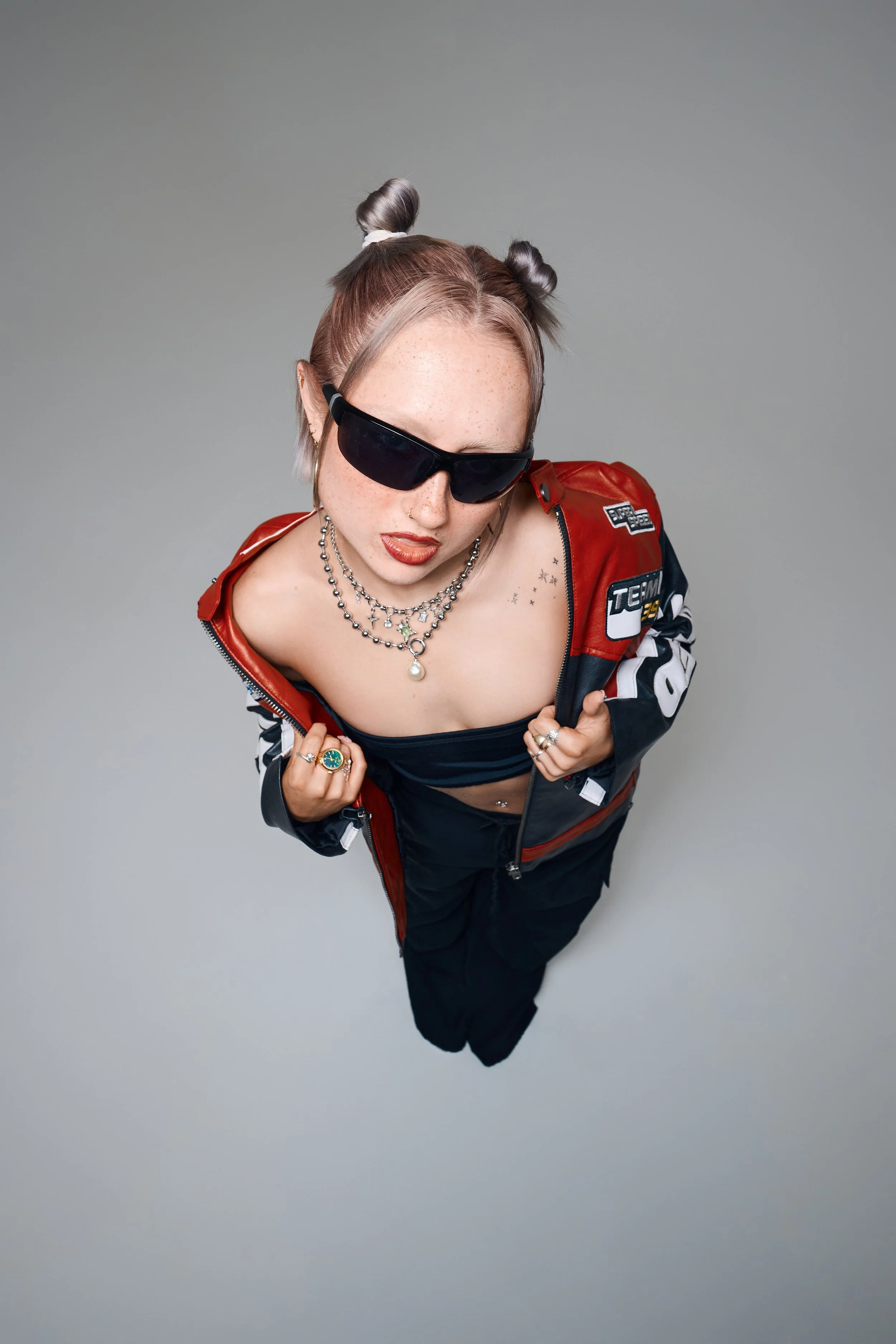 Draufsicht eines Portraits im Studio. Person trägt eine rot-schwarze Lederjacke und dunkle Sonnenbrille vor grauem Hintergrund.