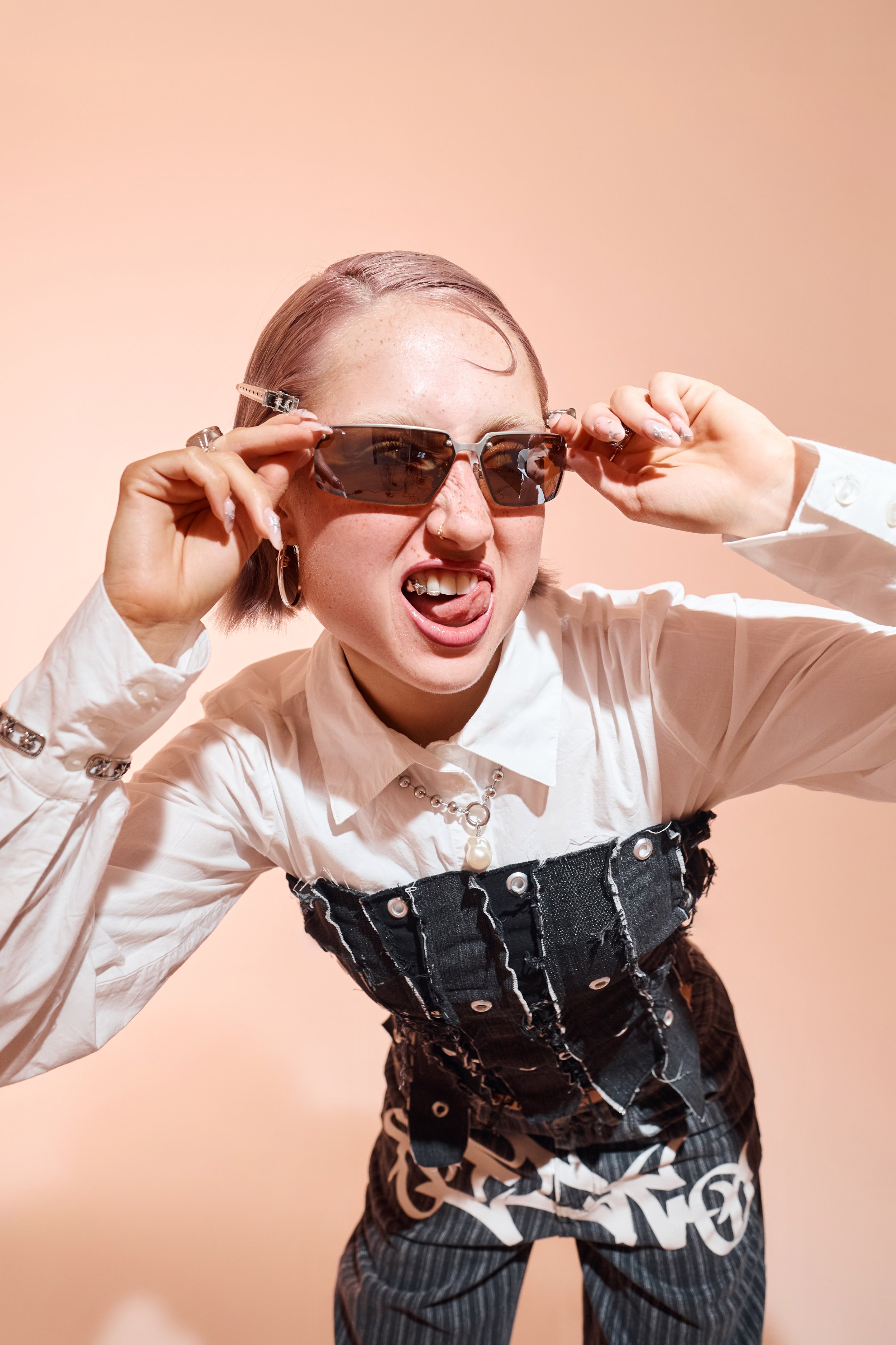Nahaufnahme vor beigem Hintergrund. Person beugt sich vor, hält eine Sonnenbrille und trägt ein Denim-Korsett über weißem Hemd.
