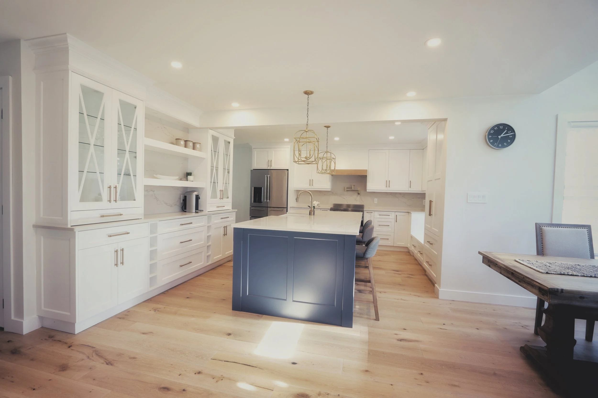 Modern kitchen with white cabinets, a navy blue kitchen island, open shelves, stainless steel appliances, and hardwood floors, illuminated by recessed lighting and pendant lights.