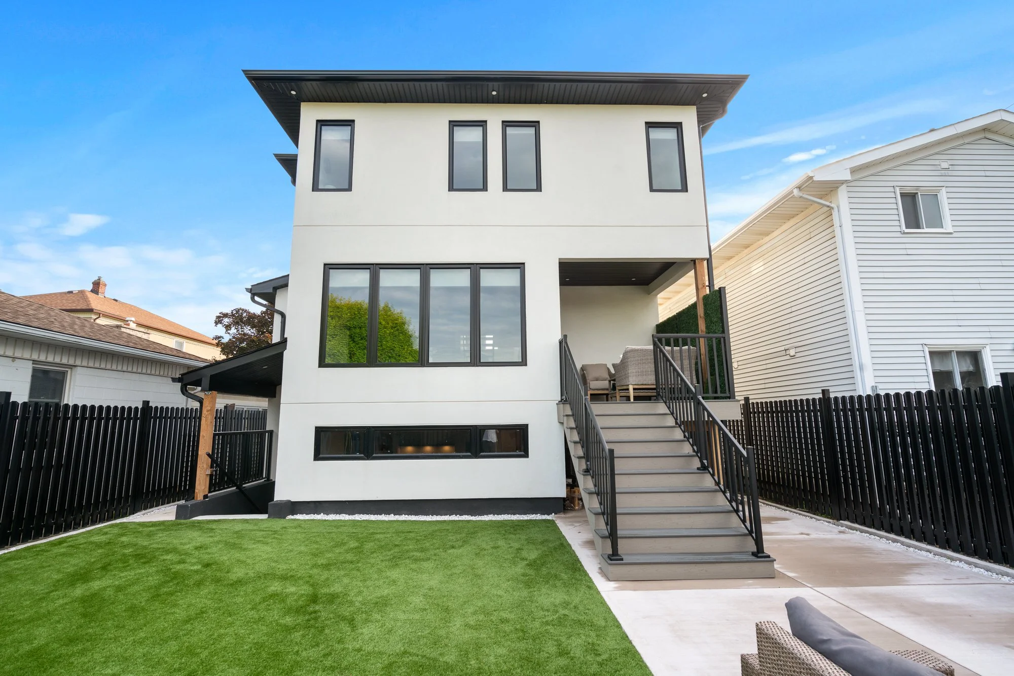 Modern white multi-story house with large windows, a black fence, a small porch, and a green lawn under a blue sky.