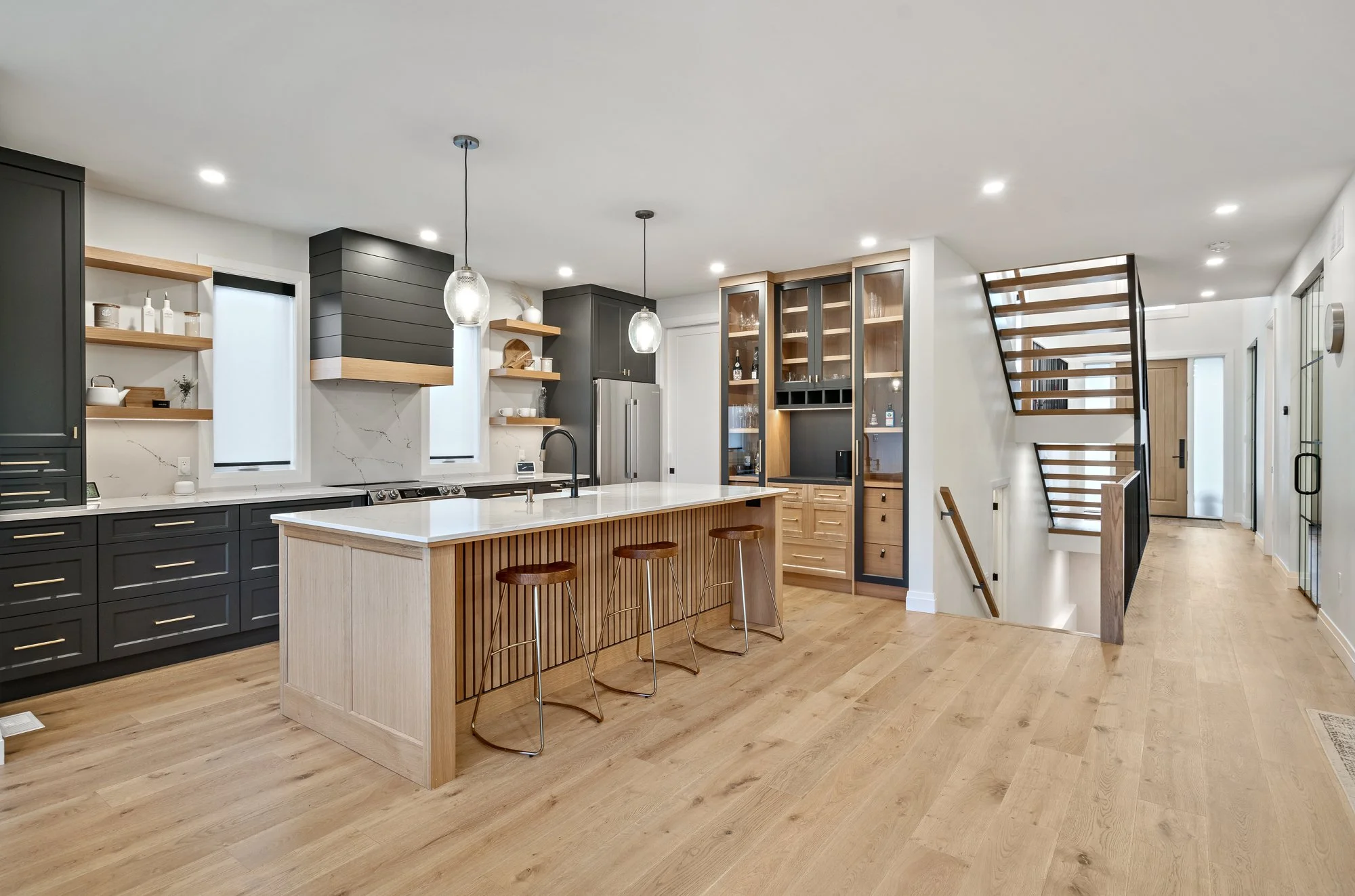 Modern kitchen with black cabinets, wooden accents, white countertops, and a kitchen island with three stools. Open shelving, stainless steel appliances, and a staircase are visible.