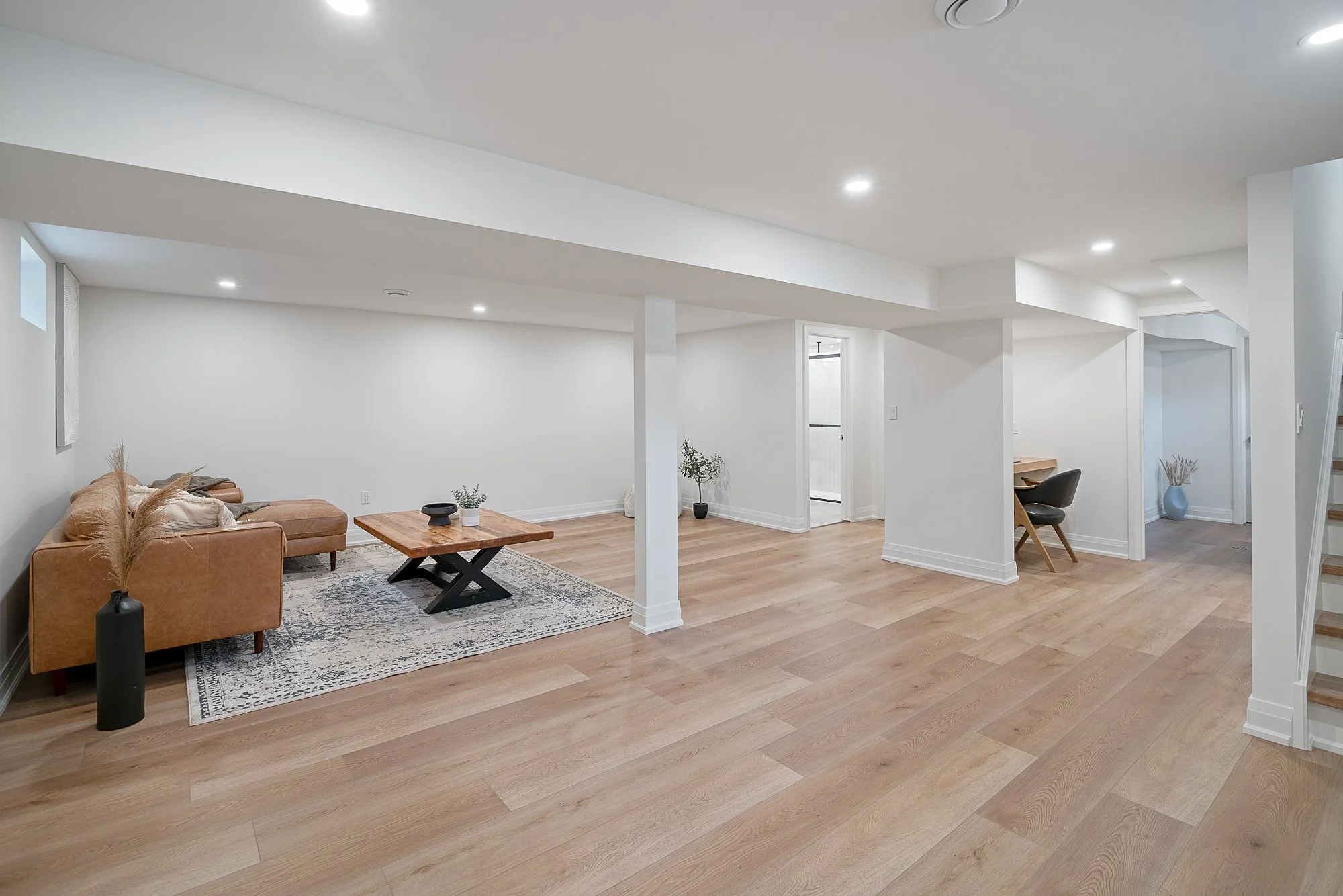 Spacious, minimally furnished basement with light wood flooring, white walls, and ceiling, containing a tan sectional sofa with pillows, a wooden coffee table, small decorative vases, plants, and a small desk area with a chair.