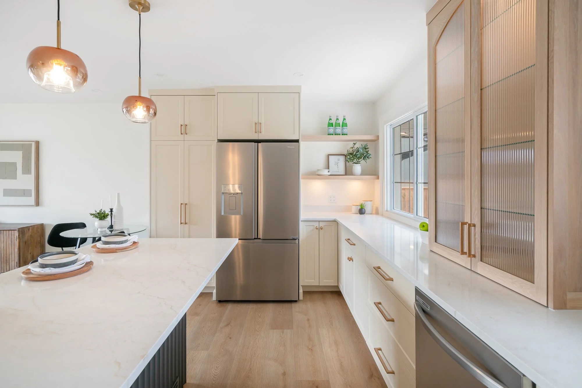 Modern kitchen with white cabinets, marble countertops, and stainless steel appliances, decorated with plants and minimalistic decor, illuminated by pendant lights.
