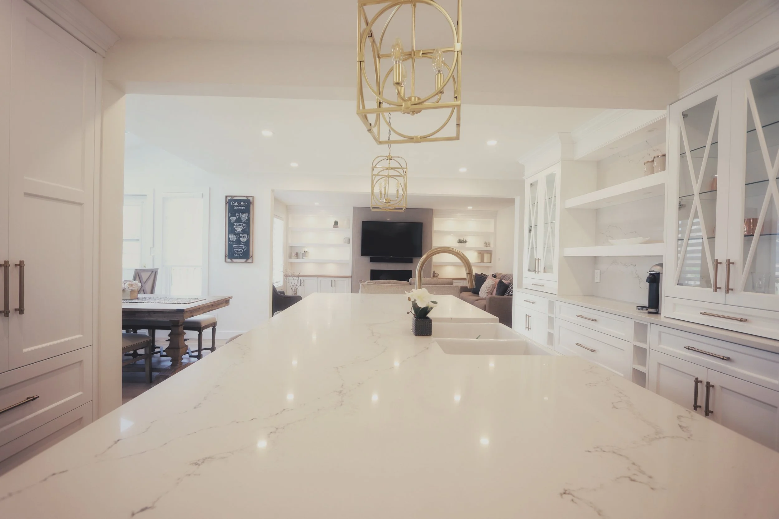 Modern kitchen with white cabinetry, marble countertop island, and a living room in the background featuring a TV and comfortable seating.