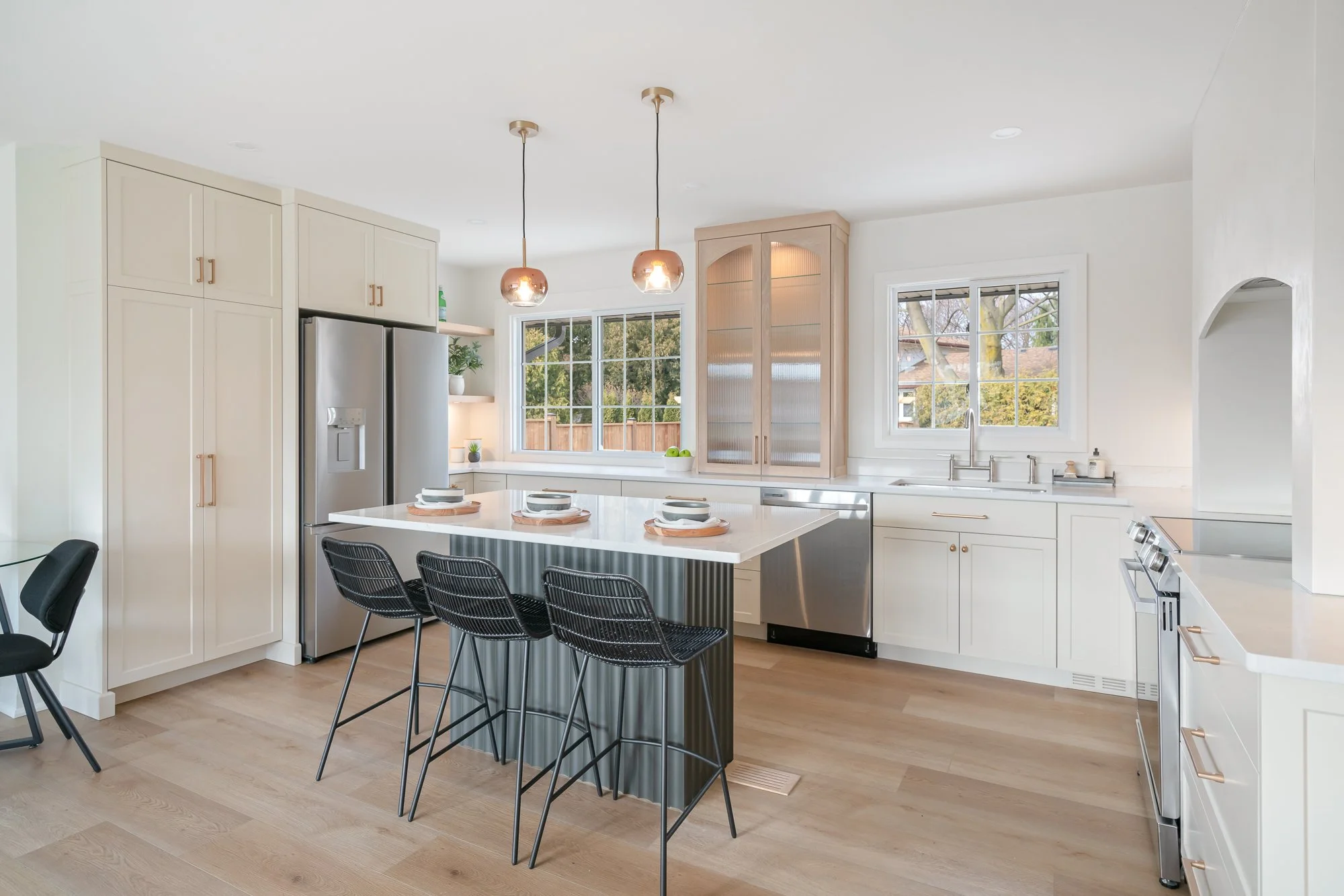 Modern kitchen with white cabinets, stainless steel appliances, a kitchen island with black chairs, large windows, and light wood flooring
