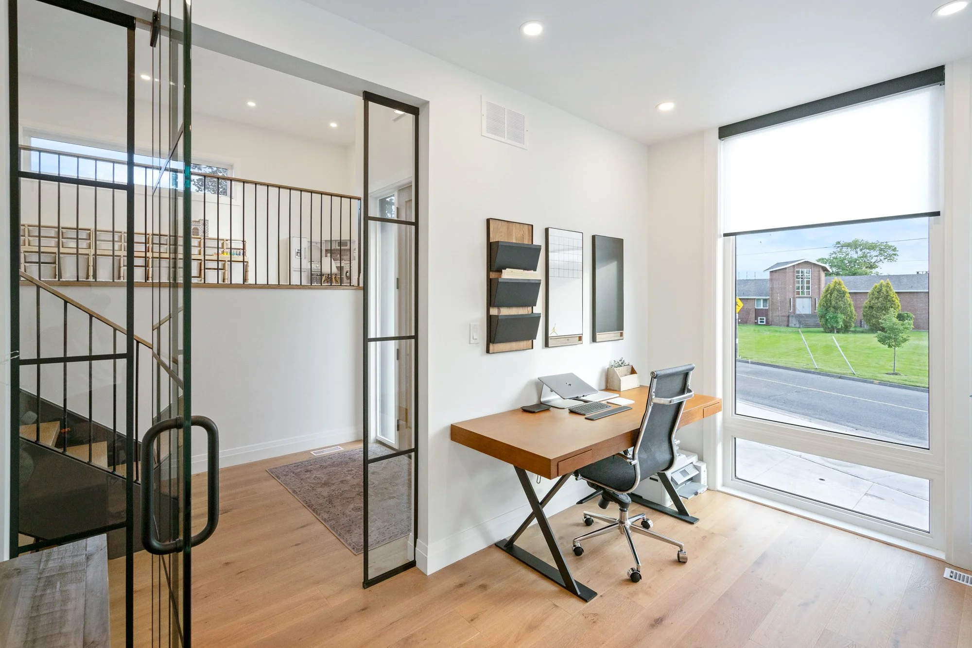 Home office with wooden desk, black ergonomic chair, large window showing neighborhood, and wall-mounted planners