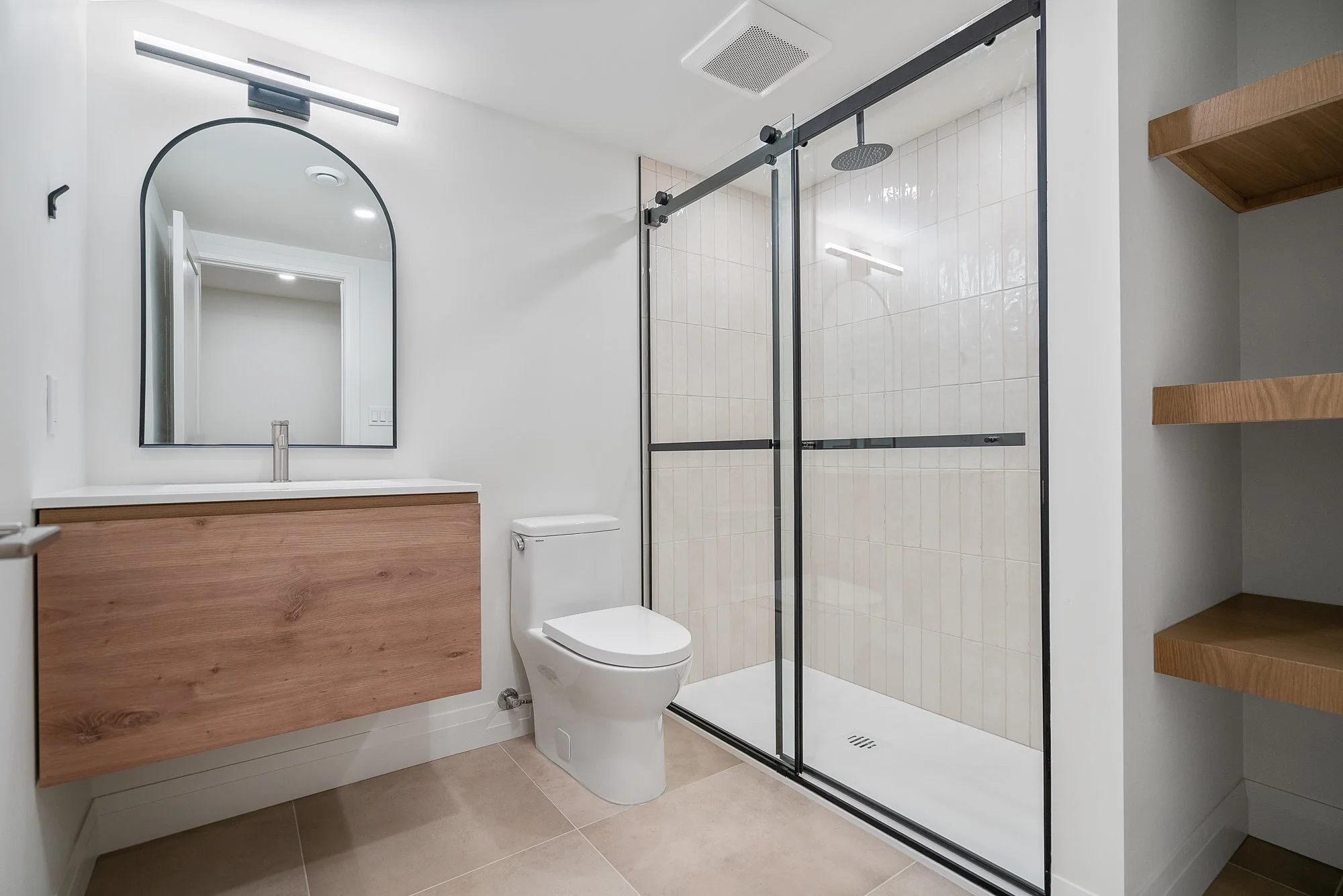 Modern bathroom with a floating wooden vanity, large mirror, glass shower enclosure with black frame, white toilet, and beige tiled floor.